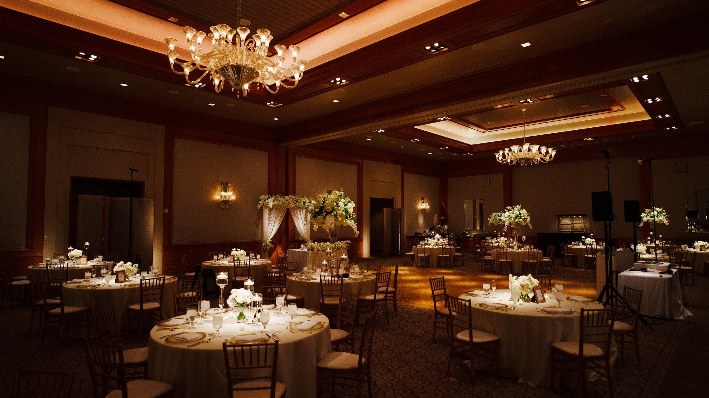 A dimly lit, elegant wedding reception hall evokes the timeless charm of a Duke Chapel ceremony, with round tables adorned with white tablecloths and floral centerpieces. Chandeliers hang from the ceiling, casting a warm glow, reminiscent of the serene beauty of Umstead's natural grace.