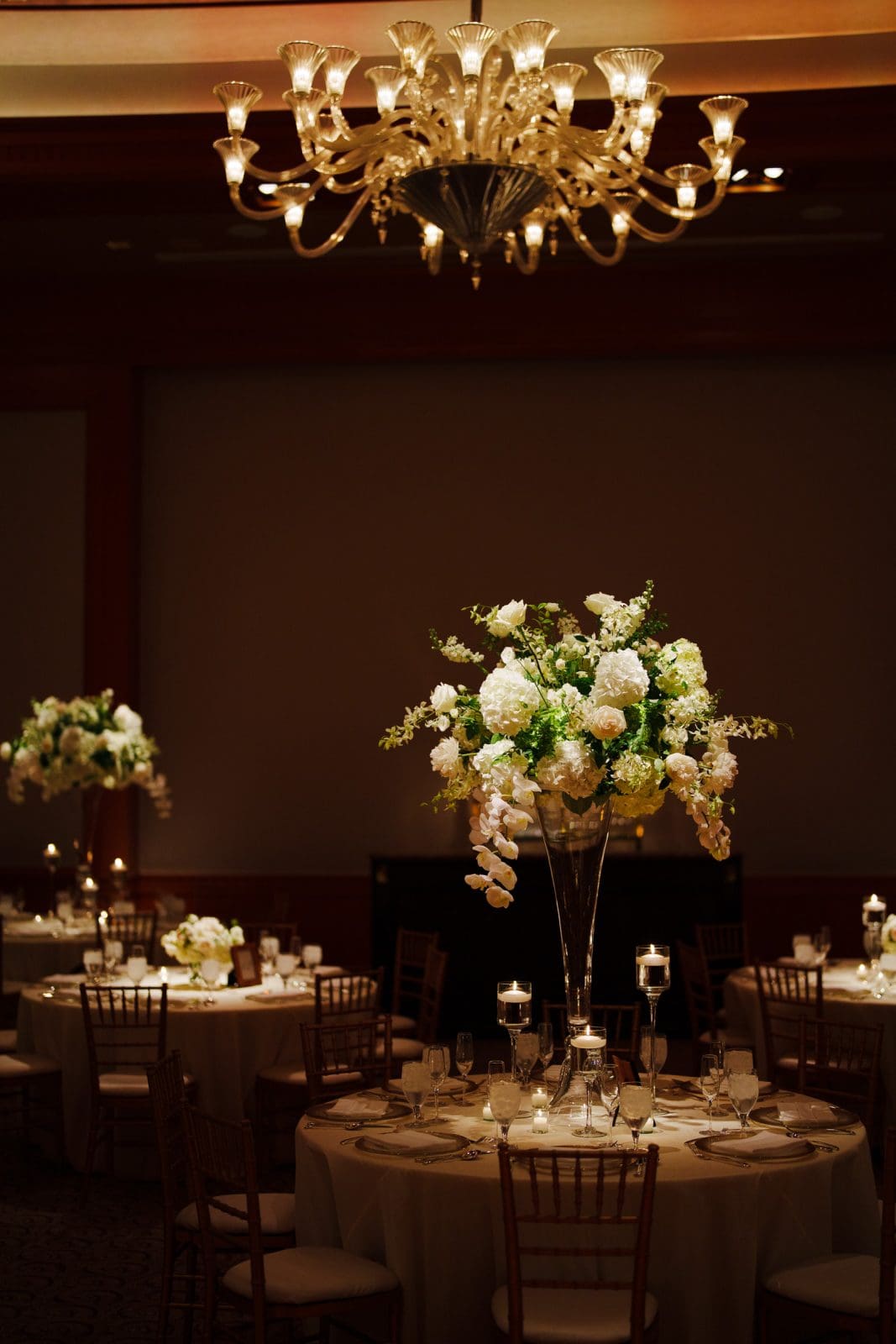 A dimly lit, elegant banquet hall reminiscent of Duke Chapel, with round tables covered in white tablecloths. Each table features tall floral centerpieces with white and green flowers. A grand chandelier hangs overhead, casting a warm glow akin to the ambiance at Umstead.