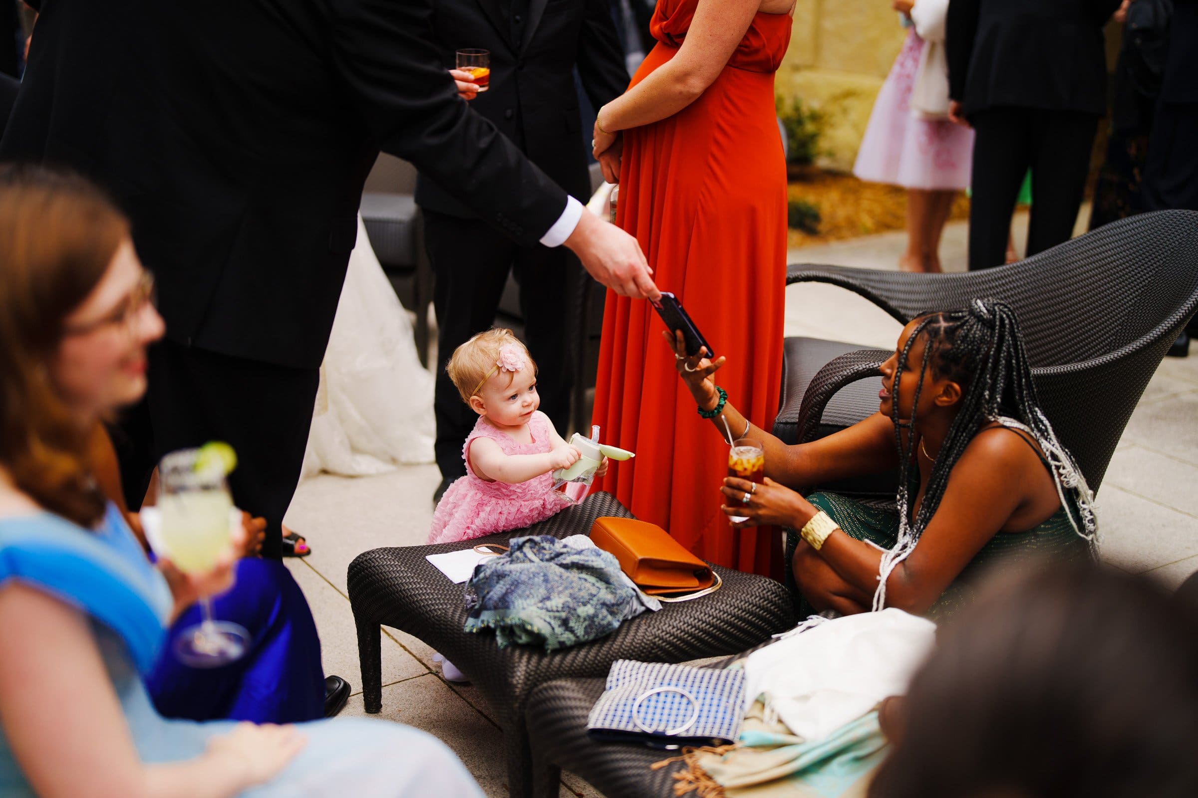 A baby in a pink dress smiles and reaches for a phone held by someone in a black suit near Duke Chapel. A seated woman with braided hair is handing something to the baby. Other people with drinks are around, creating a lively social scene, reminiscent of gatherings at Umstead.