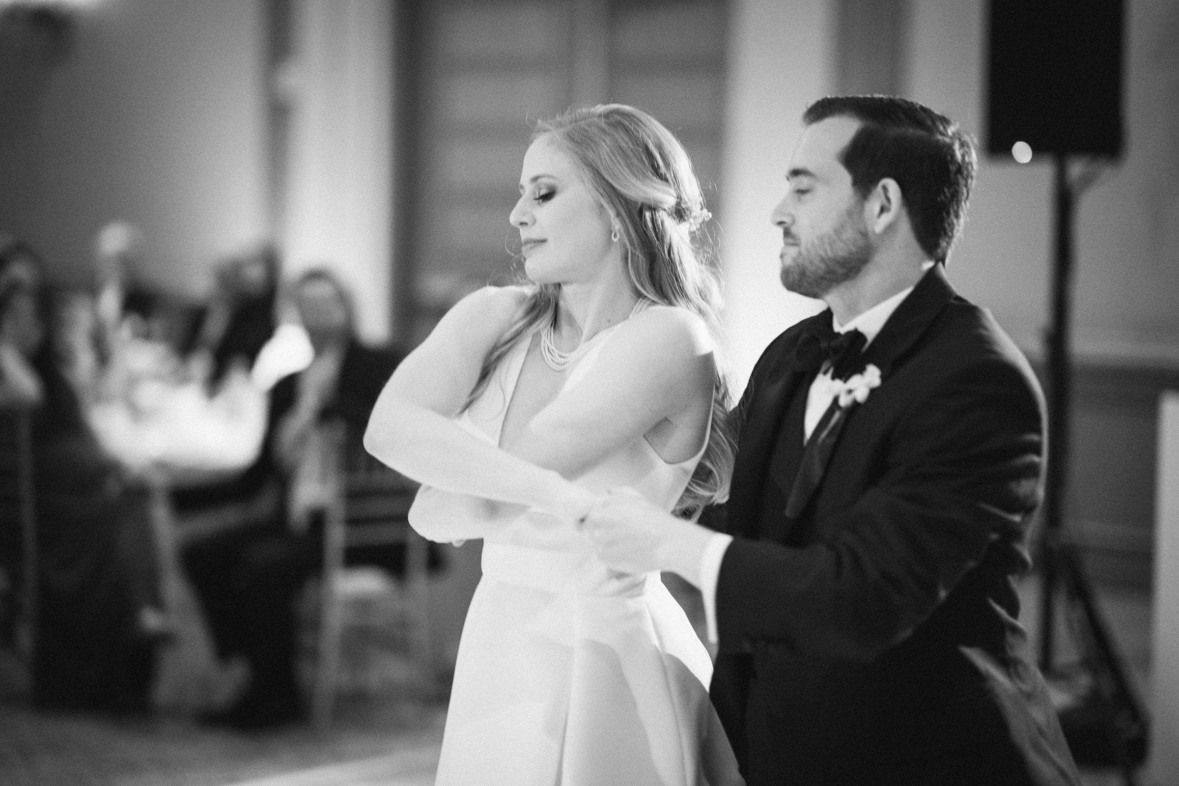A black and white photo captures a couple gracefully dancing at what could be a formal event near Duke Chapel. The woman is in an elegant dress, and the man, wearing a suit and boutonniere, complements her beautifully. Softly focused tables with guests seated add depth to this timeless Umstead-like scene.