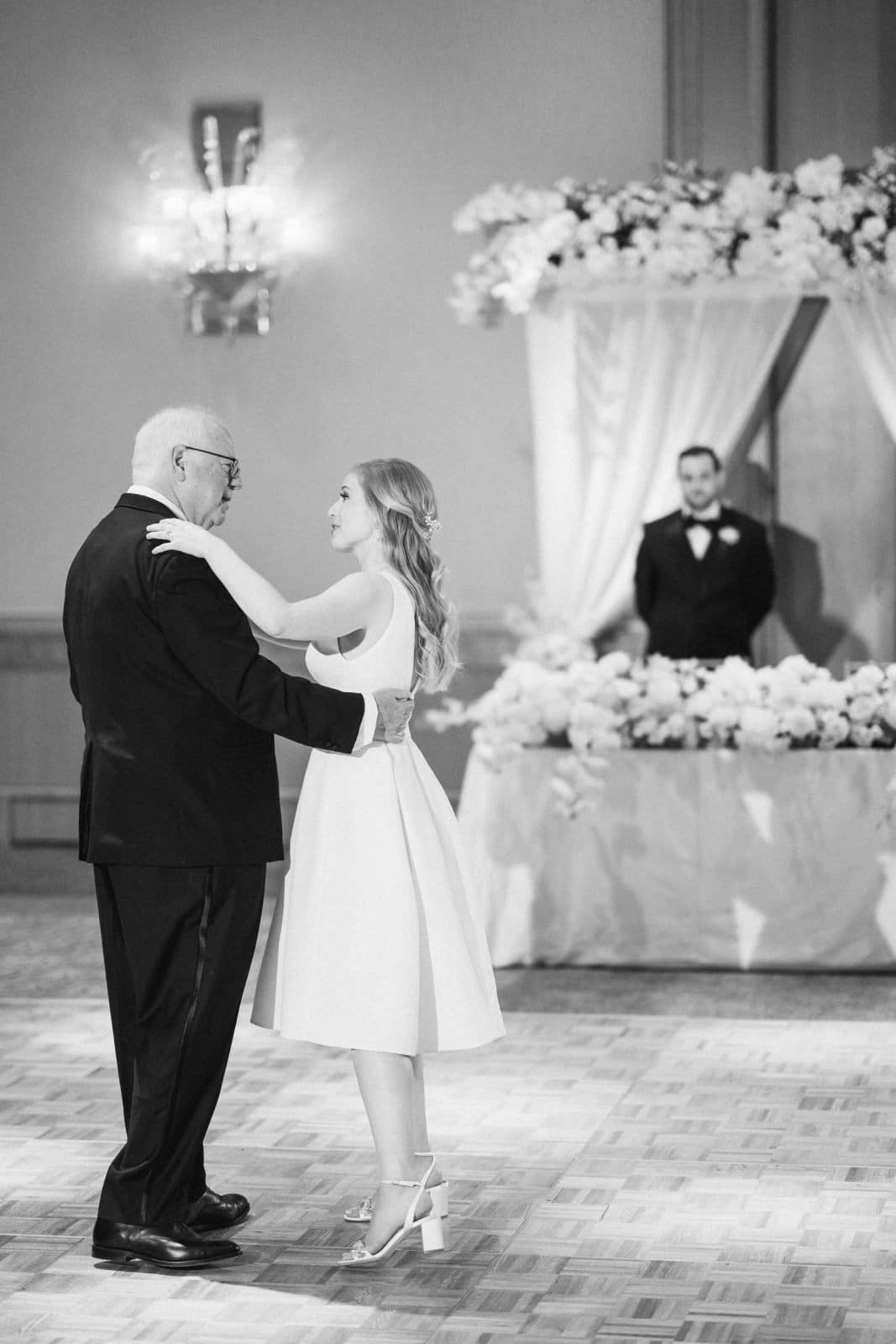 In a black-and-white photo, a couple dances with elegance; the man in a suit and the woman in a sleeveless dress. They glide gracefully through a decorated room, where floral arrangements echo the refinement of an event at Duke Chapel, as another tuxedo-clad man stands by a table.