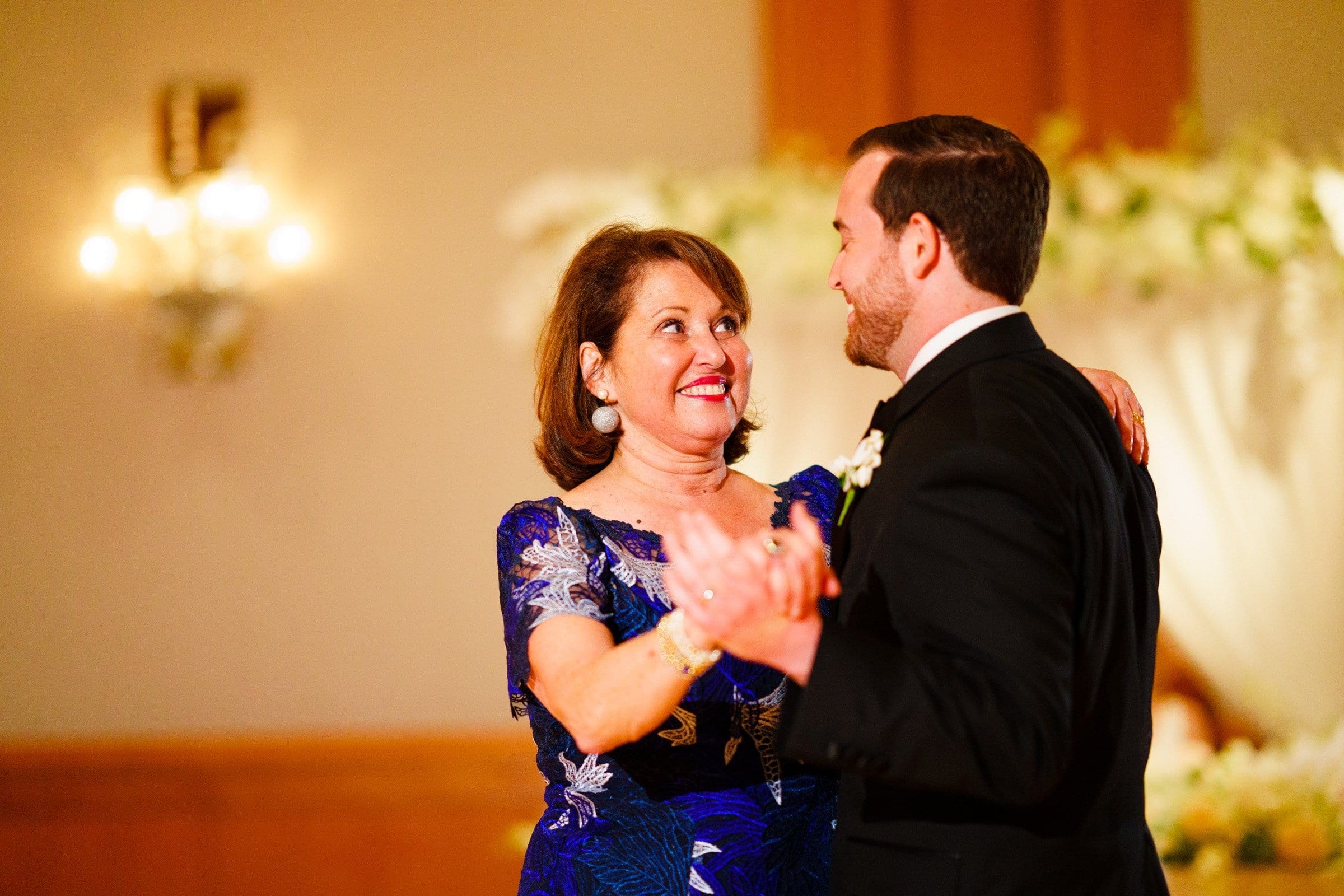 A woman in a blue dress and a man in a black suit dance together at Duke Chapel, both smiling warmly. The background features soft lighting and floral decorations from Umstead, suggesting a celebratory event.