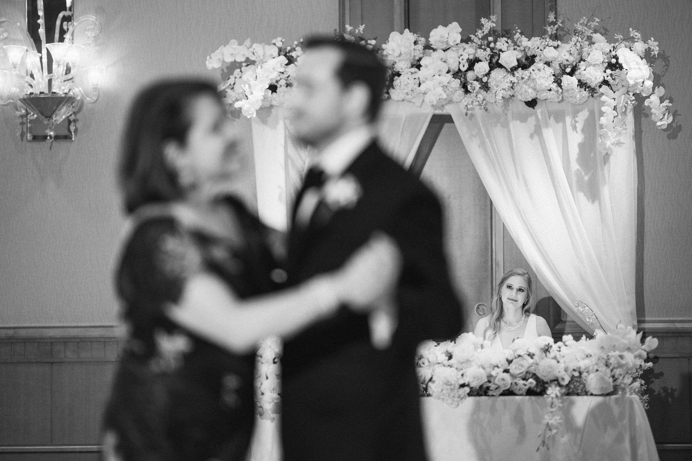 A black and white image captures a couple dancing gracefully in the foreground, while in the background, a woman sits at a flower-adorned table. The elegant setting evokes the charm of a venue like Duke Chapel or Umstead.