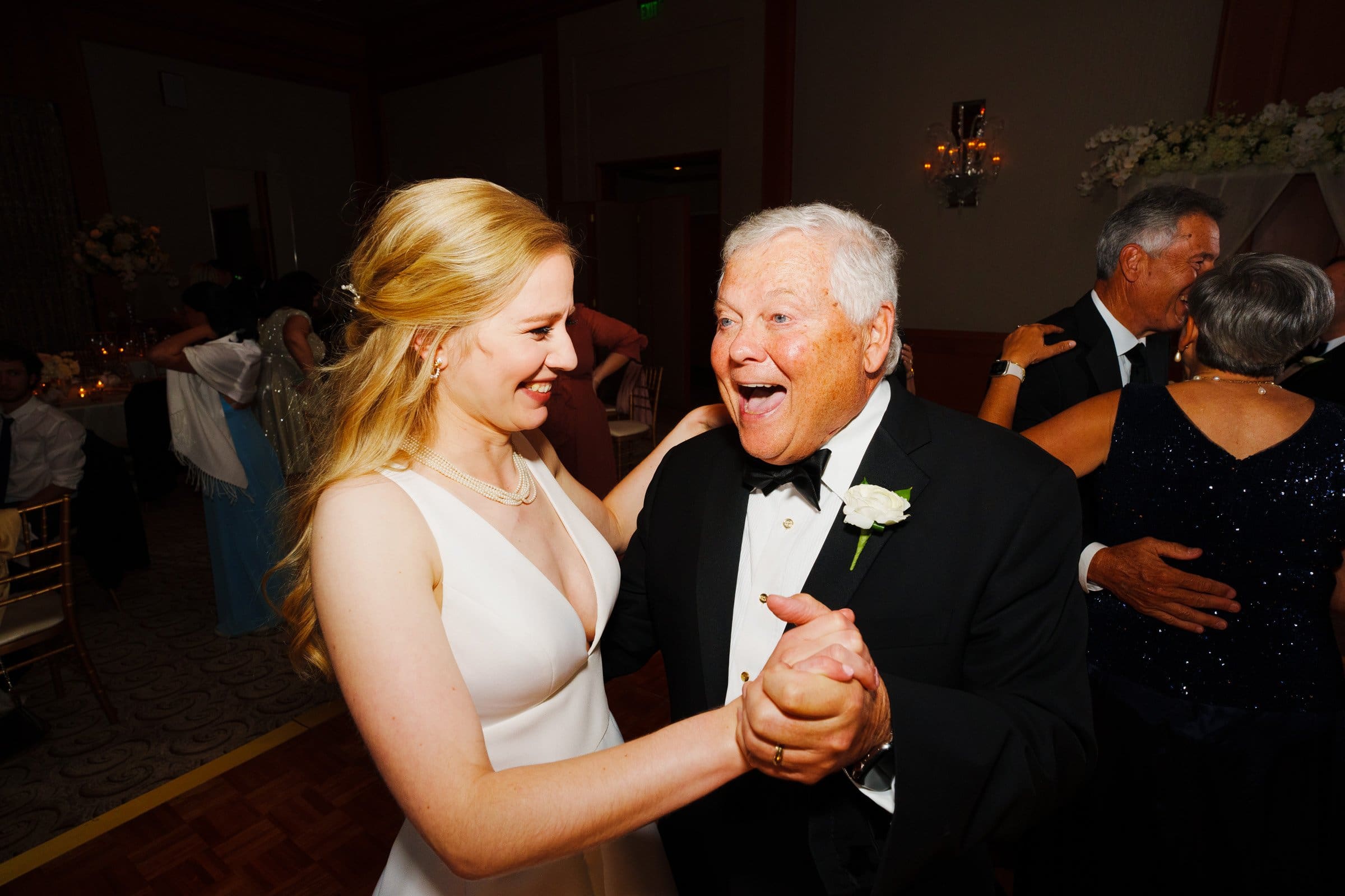 A young woman in a white dress and an elderly man in a black tuxedo joyfully dance together at an elegant indoor event at Umstead. Both are smiling warmly, surrounded by other couples dancing. The atmosphere is festive and bright, reminiscent of celebrations at Duke Chapel.