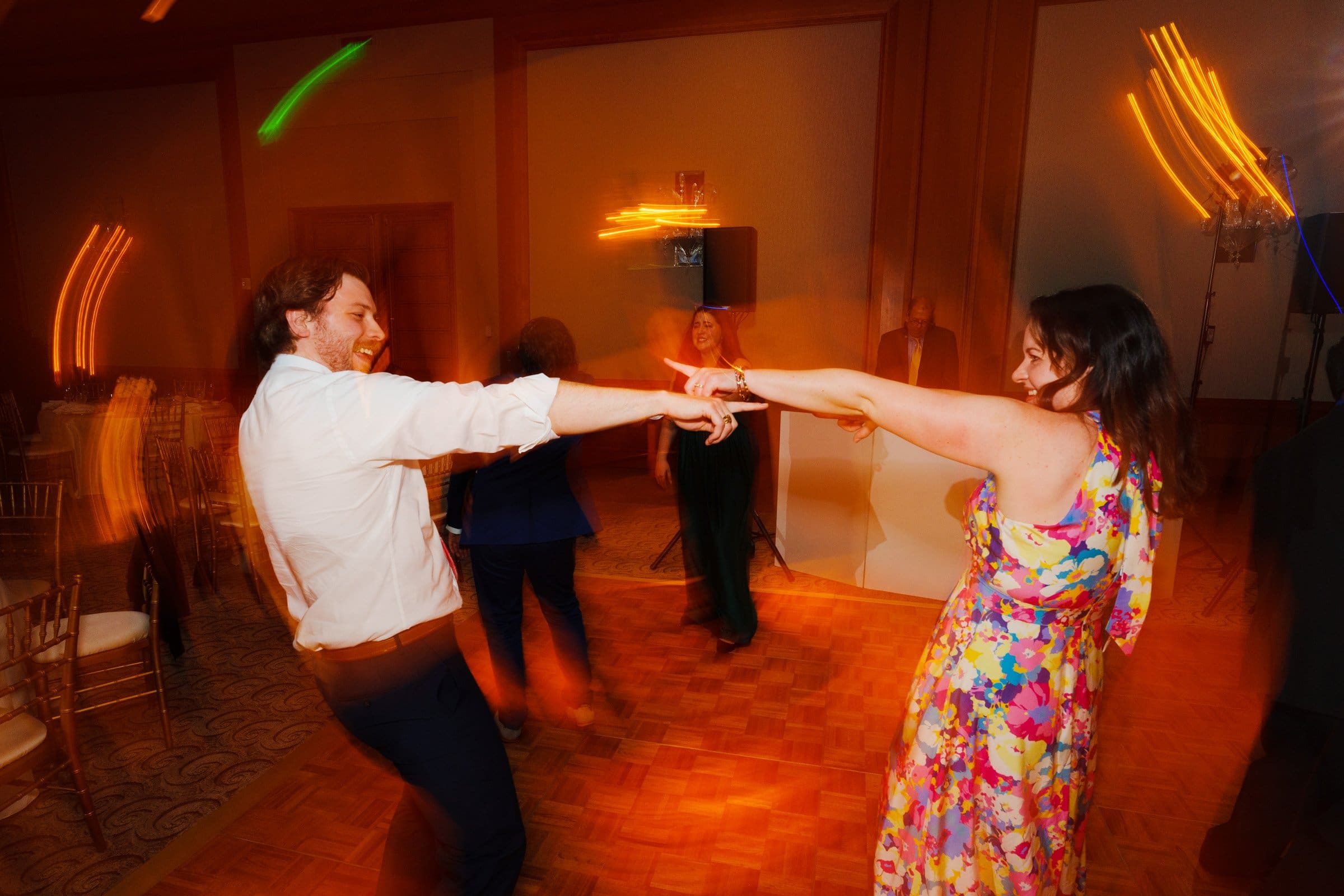 A man and woman dance energetically in a warmly lit room with a wooden floor reminiscent of Duke Chapel's warmth. She wears a colorful floral dress, and he has on a white shirt and dark pants. The lights blur, creating a lively atmosphere as if echoing the serenity of Umstead. Two people and a DJ booth are in the background.