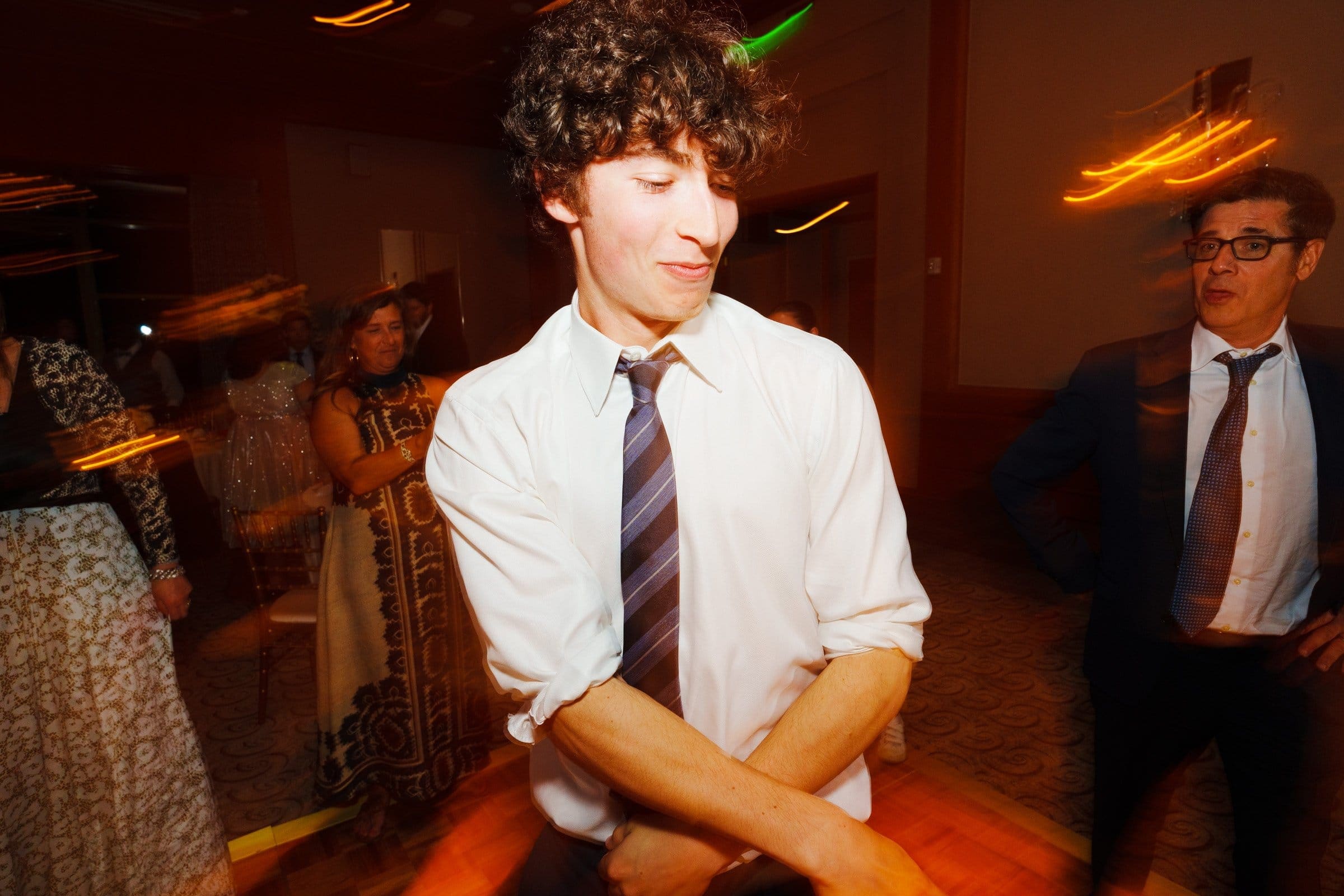 A man with curly hair dances energetically at a party, wearing a white shirt and striped tie. The lively atmosphere, resembling the energy of a night near Duke Chapel, is enhanced by blurred motion lights. Other guests, including a man in glasses and a woman in a patterned dress, are visible in the background.
