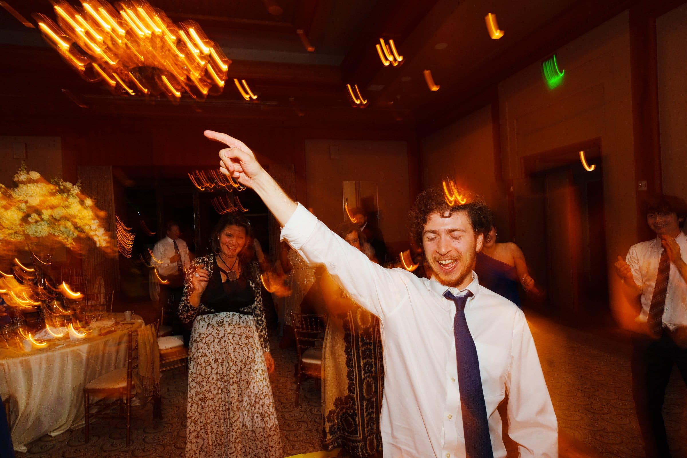 A man in a white shirt and tie dances joyfully at an indoor party, reminiscent of celebrations near Duke Chapel, with blurred lights and motion around him. A chandelier and flower arrangement are in the background, enhancing the festive atmosphere.
