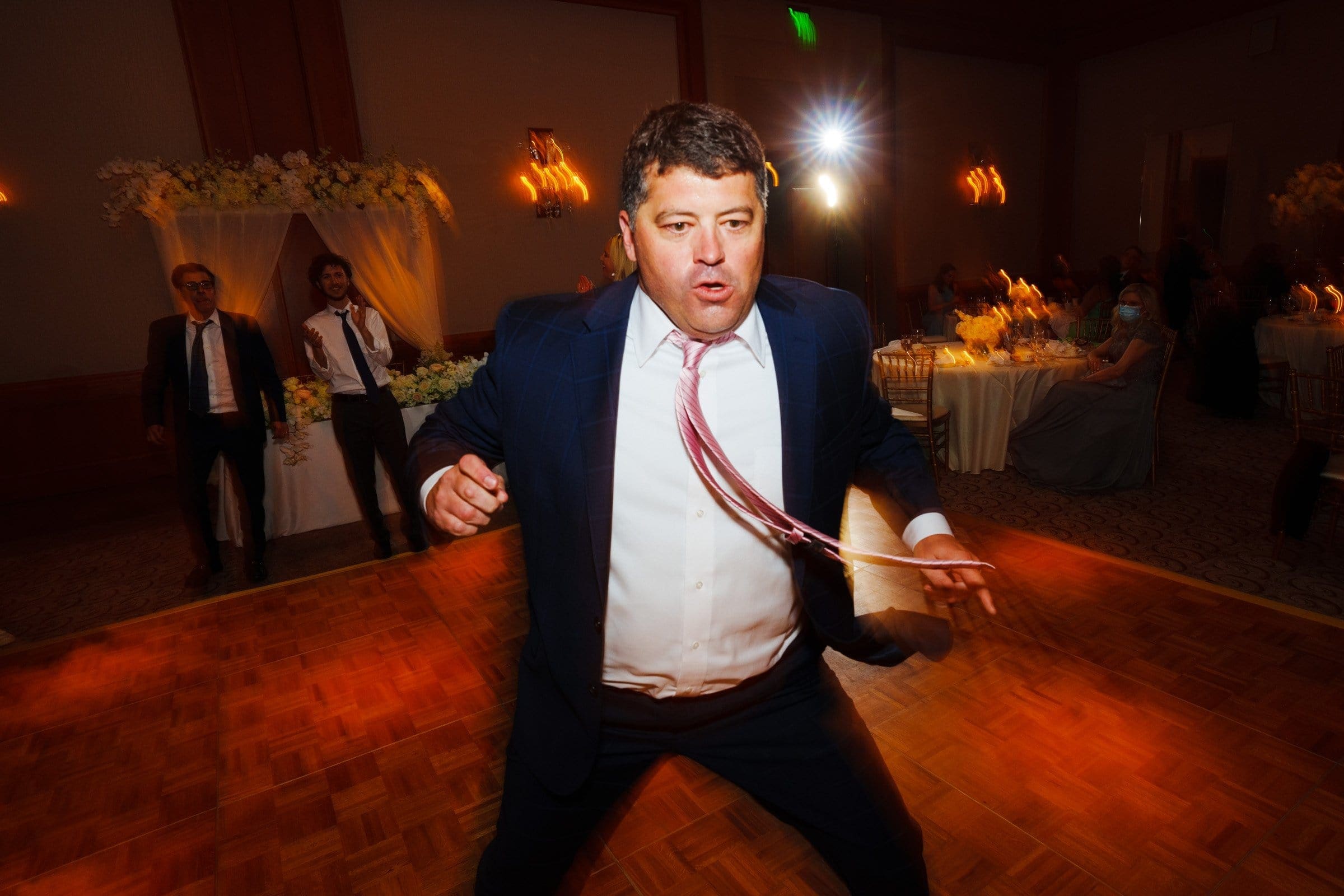 A man in a suit enthusiastically dances on the wooden floor at a lively indoor event, reminiscent of celebrations at Duke Chapel. His tie is loosened, and he seems to be reveling in the moment. In the background, guests seated at tables are visible, along with floral decorations and soft Umstead lighting.