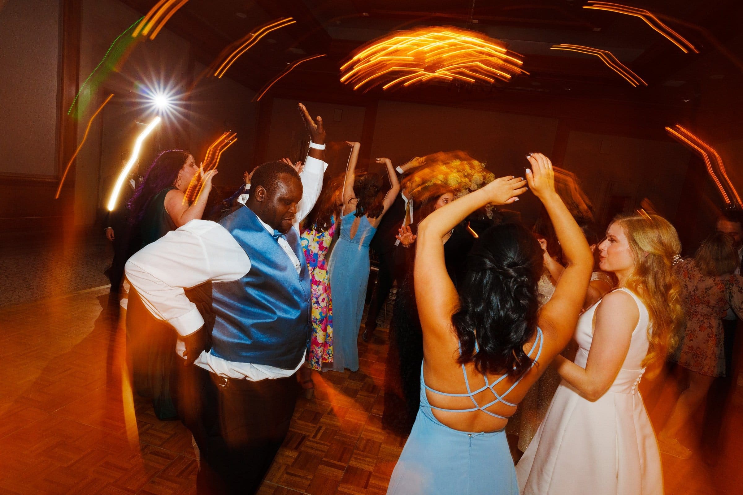 People are dancing energetically at a party near Duke Chapel, with blurred lights creating a dynamic atmosphere. A man in a blue vest and tie raises his arms, surrounded by women in colorful dresses, all enjoying the lively scene reminiscent of Umstead's vibrant surroundings.