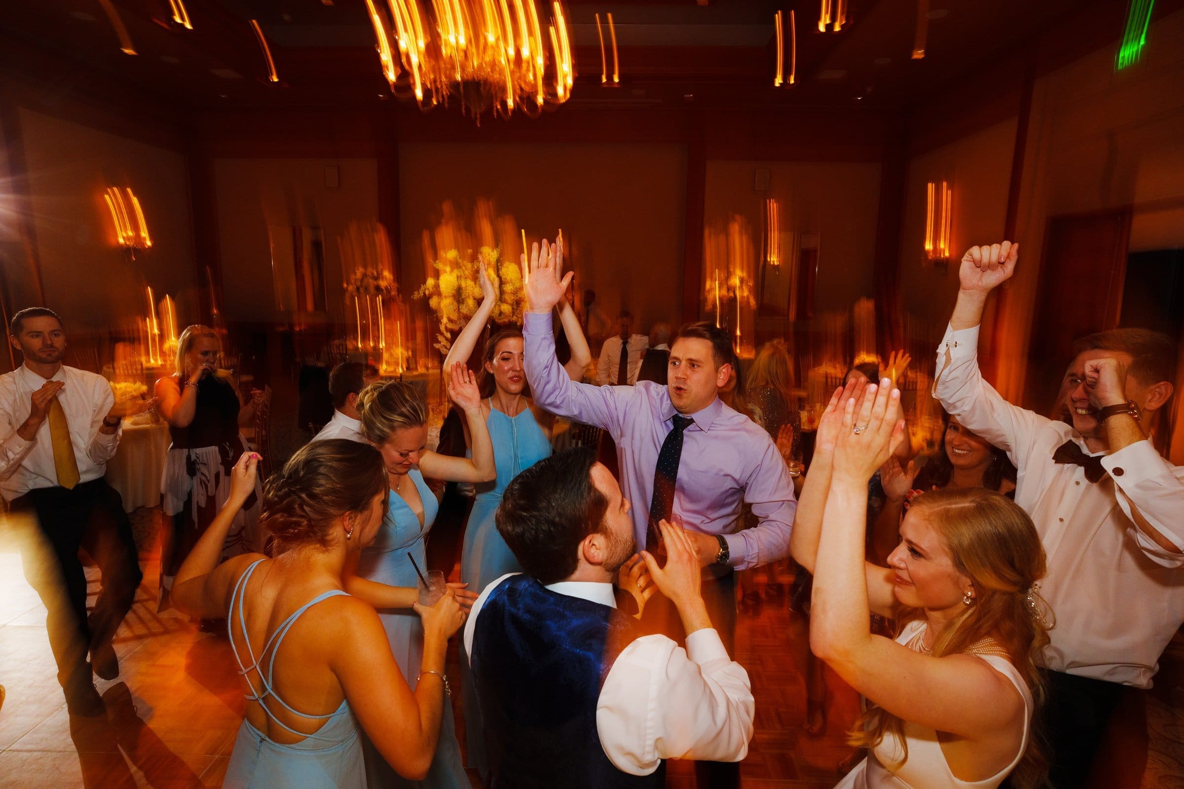A lively wedding reception unfolds beneath a grand chandelier, with guests dancing energetically. Women in light blue dresses and men in suits grace the floor. The warm, blurry lighting evokes the elegance of Duke Chapel against the vibrant backdrop, reminiscent of an evening at Umstead.