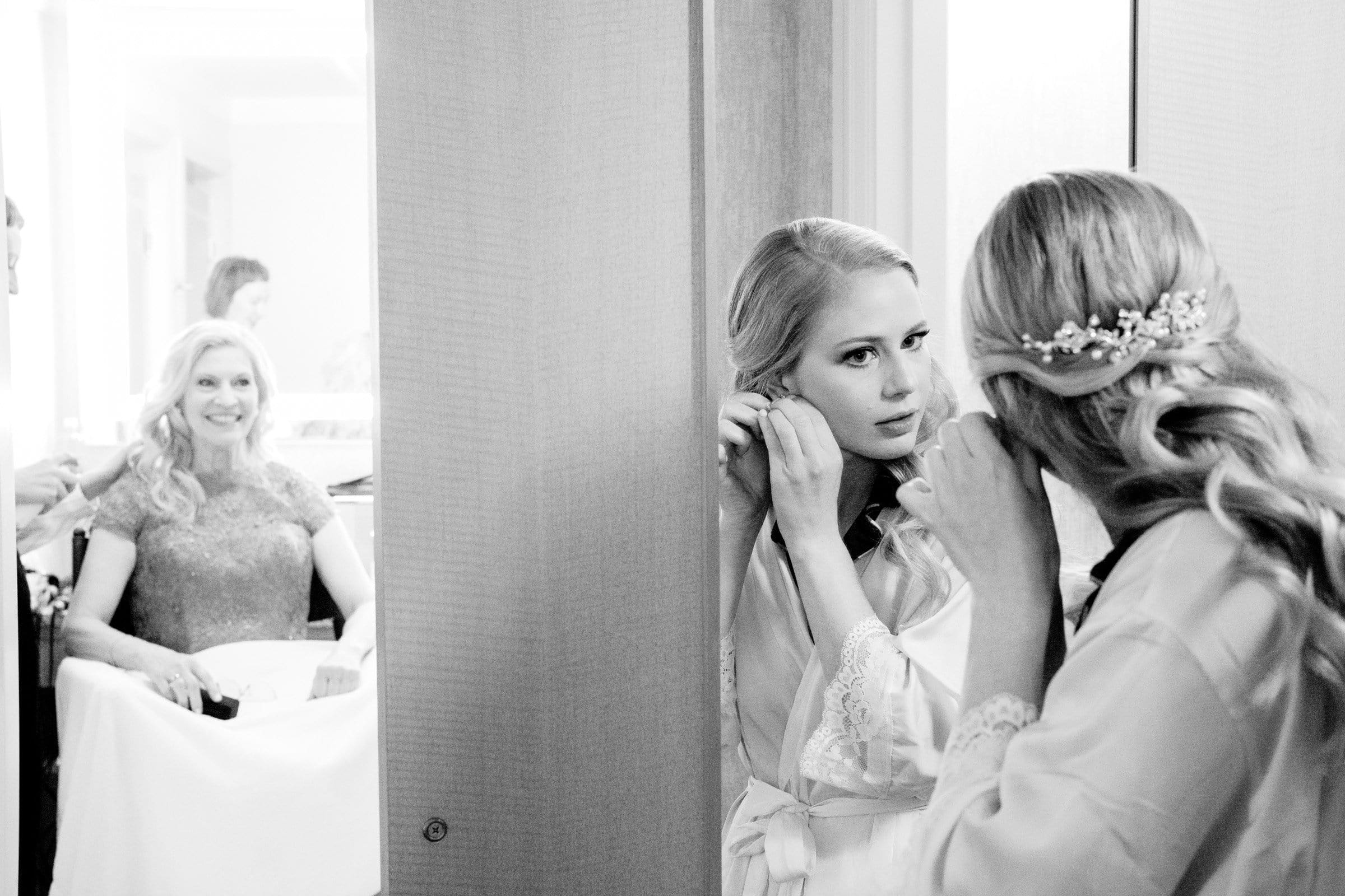 A bride adjusts her earring in a mirror, adorned with a decorative hairpiece at the enchanting Duke Chapel. In the background, a woman smiles while seated, and another person can be seen further back. The scene captures a joyful pre-wedding preparation moment at this iconic location.