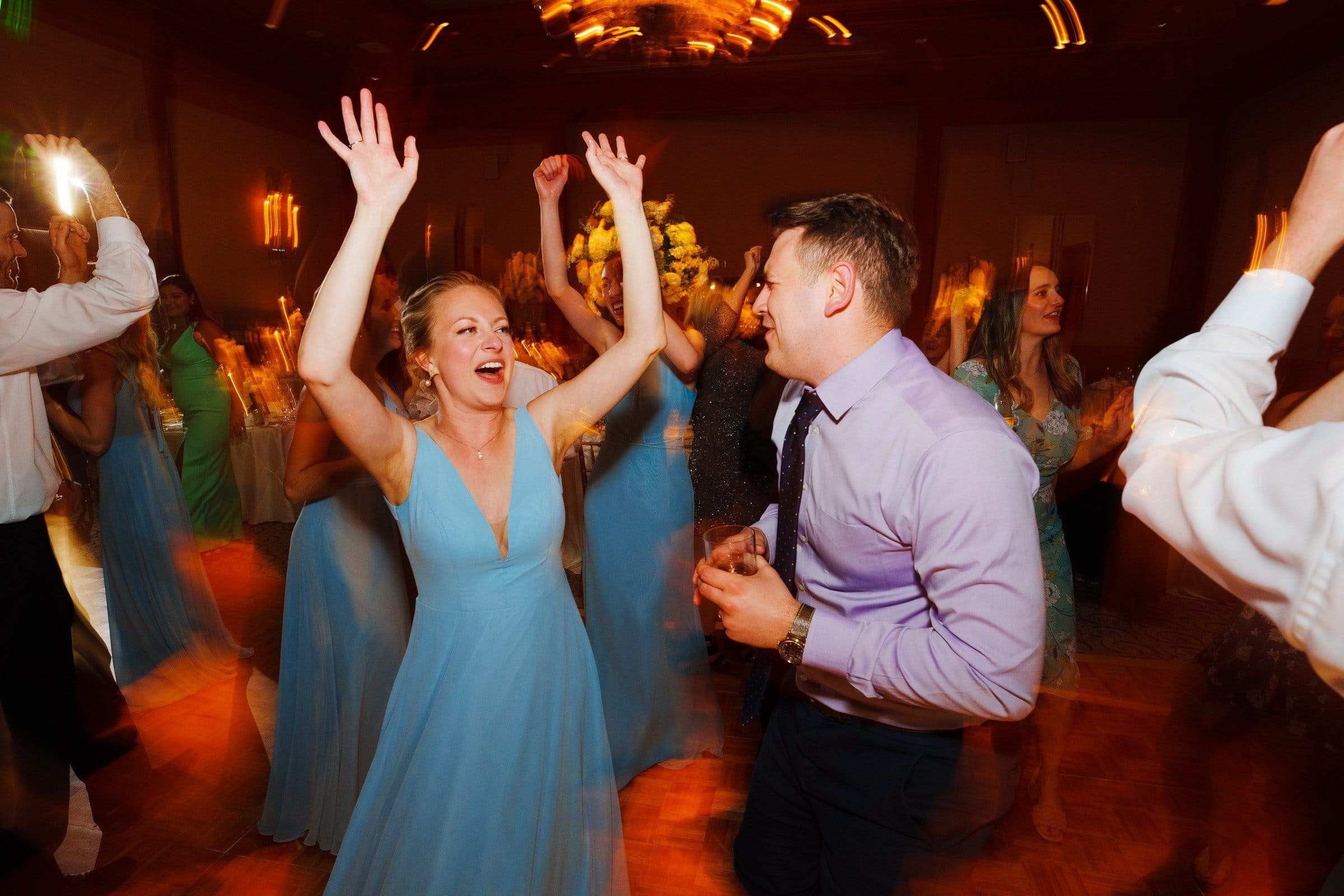 At a lively event near Duke Chapel, a woman in a blue dress and a man in a light purple shirt dance joyfully. The room is warmly lit, with others swaying in the background amid elegant floral decorations reminiscent of Umstead's natural beauty.