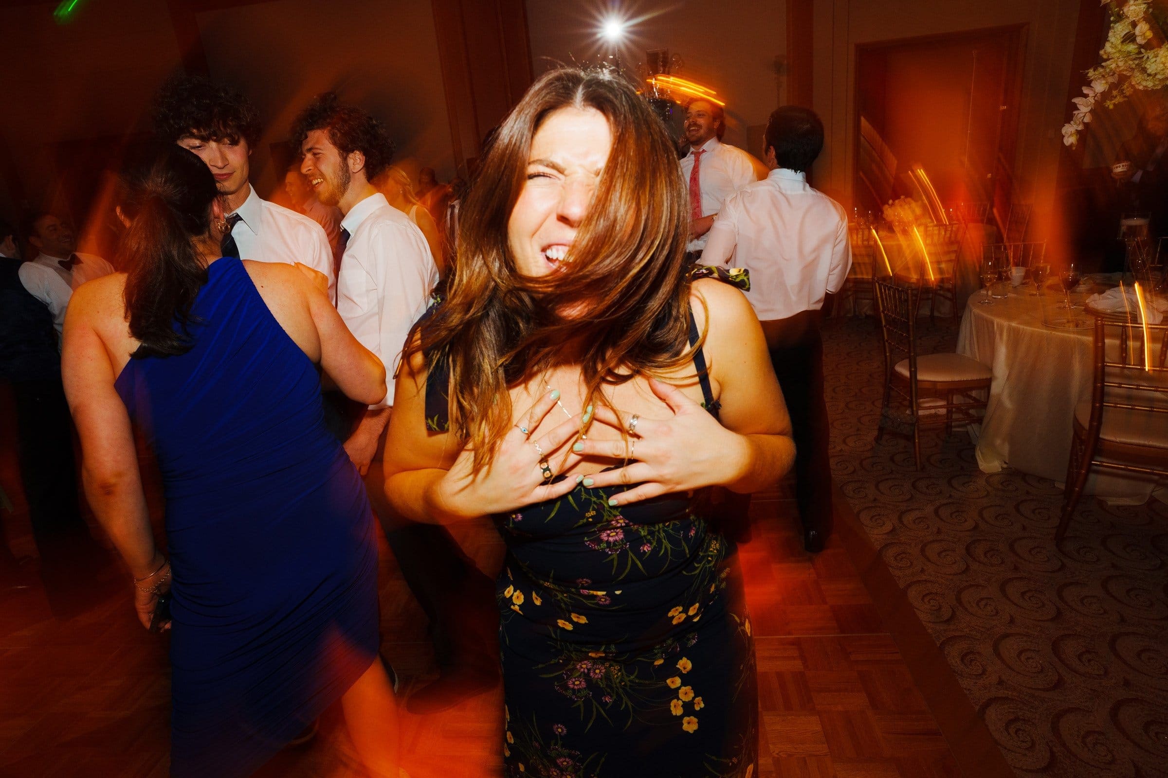 A woman in a floral dress smiles energetically while dancing at a lively party, as if celebrating alongside the vibrant spirit found near Duke Chapel. Her hair moves with the motion, and vibrant, blurred orange lights create a dynamic background. Other attendees can be seen enjoying the festive atmosphere.