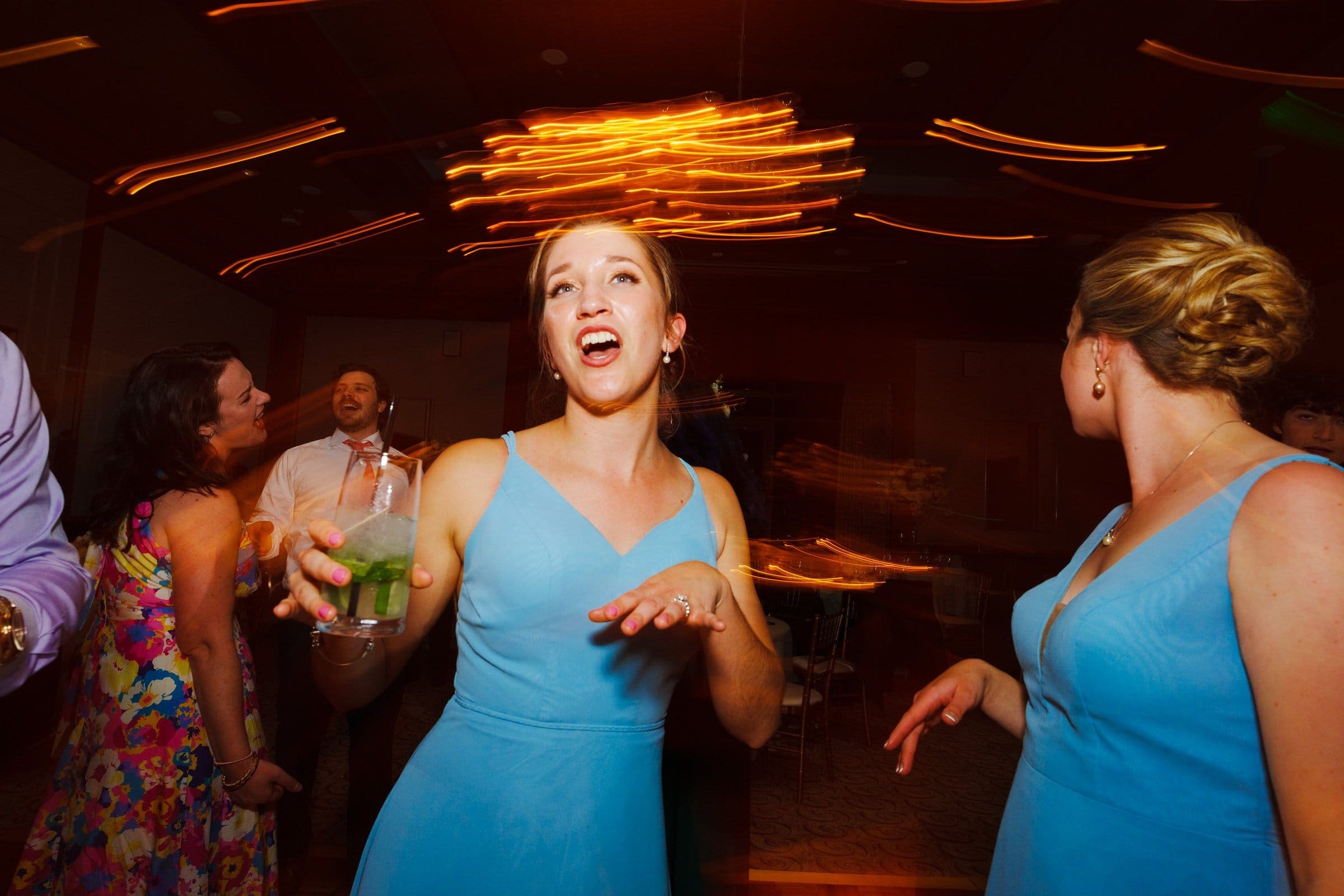 A woman in a blue dress joyfully sings while holding a drink, surrounded by others in formal attire. The lively scene, reminiscent of celebrations at Duke Chapel, features swirling light trails suggesting movement and energy.