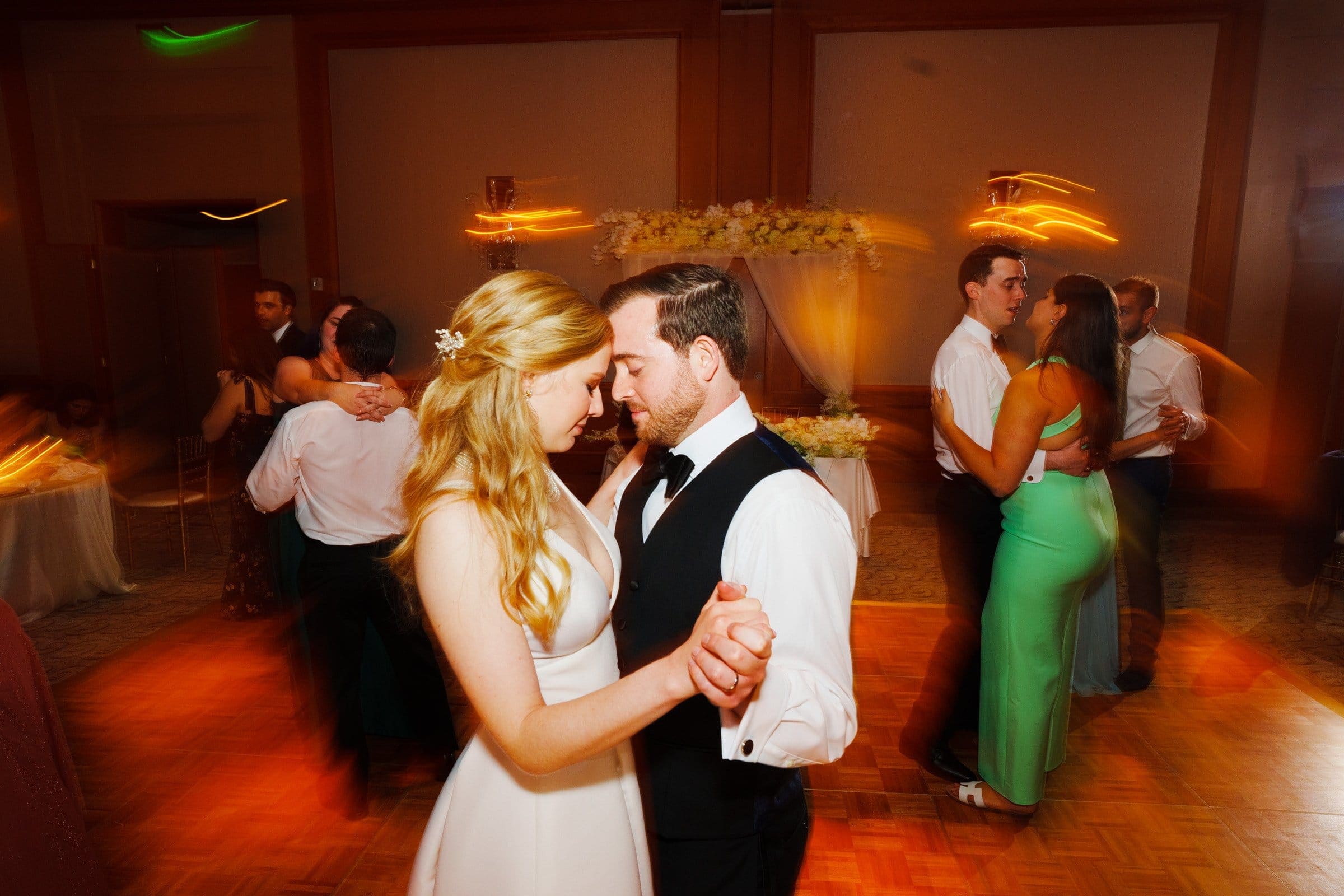A couple dances gracefully in the foreground of a warmly lit reception hall reminiscent of Duke Chapel's grandeur. The woman wears a white dress, and the man wears a suit with a vest. Blurred figures sway in the background under soft Umstead-like lighting.