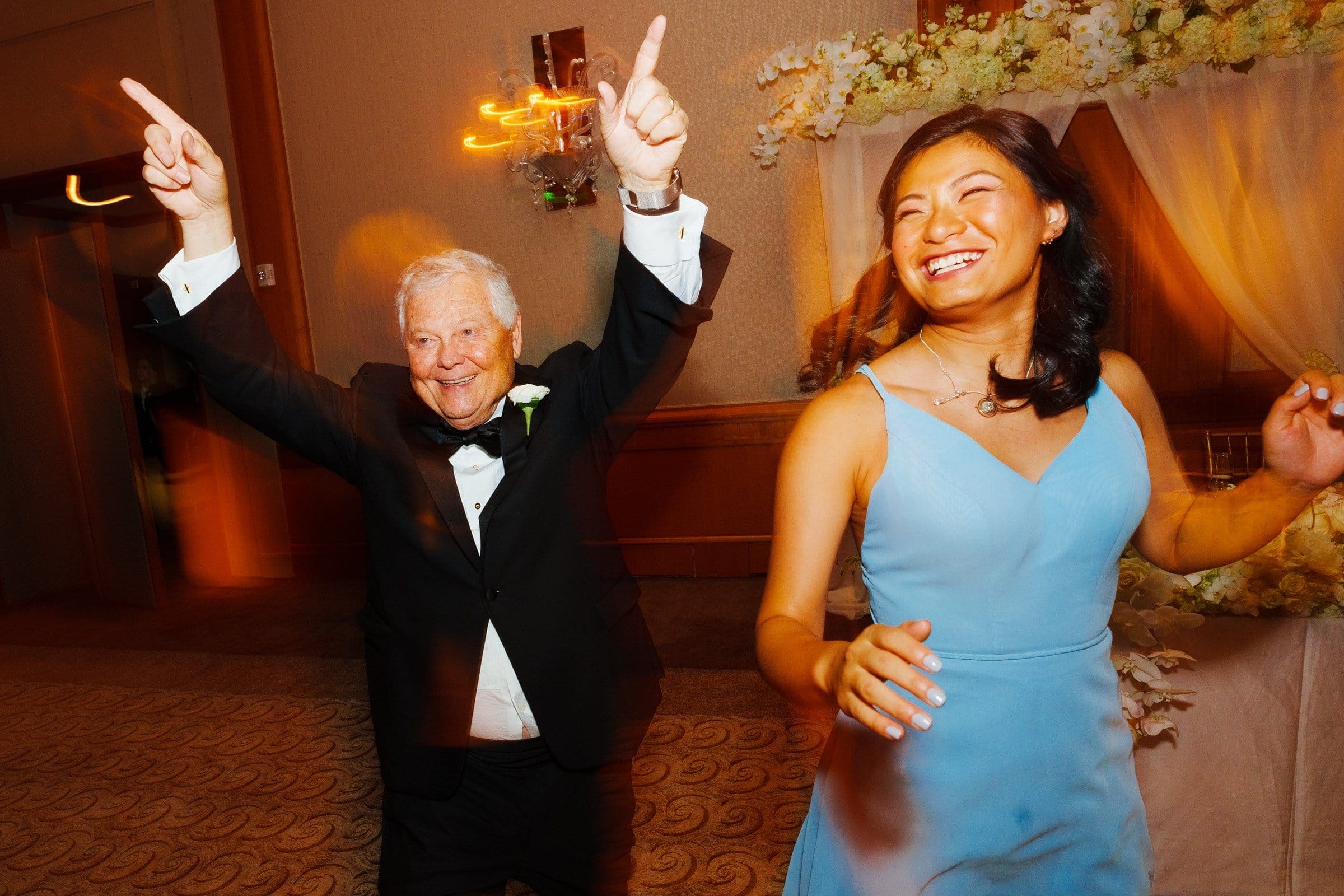 An elderly man in a tuxedo and a young woman in a blue dress joyfully dance at a formal event held at Duke Chapel. The background features floral decorations and warm lighting, creating a festive atmosphere reminiscent of evenings spent at Umstead.