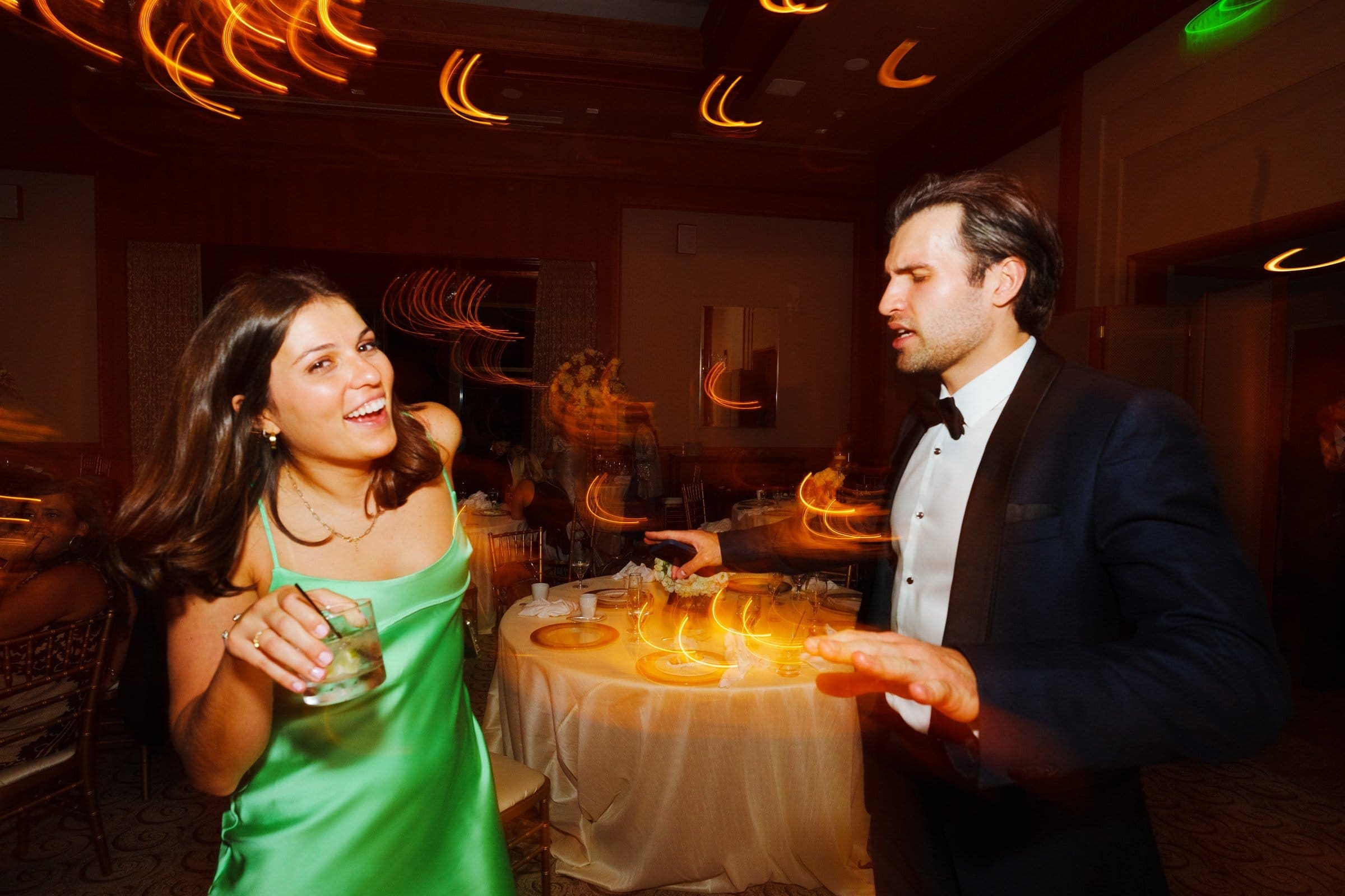 A woman in a green dress laughs and holds a drink while a man in a tuxedo dances, reminiscent of an elegant evening at Duke Chapel. They are in a lively room with blurred motion lights and a round table, capturing the festive atmosphere akin to celebrations at Umstead.