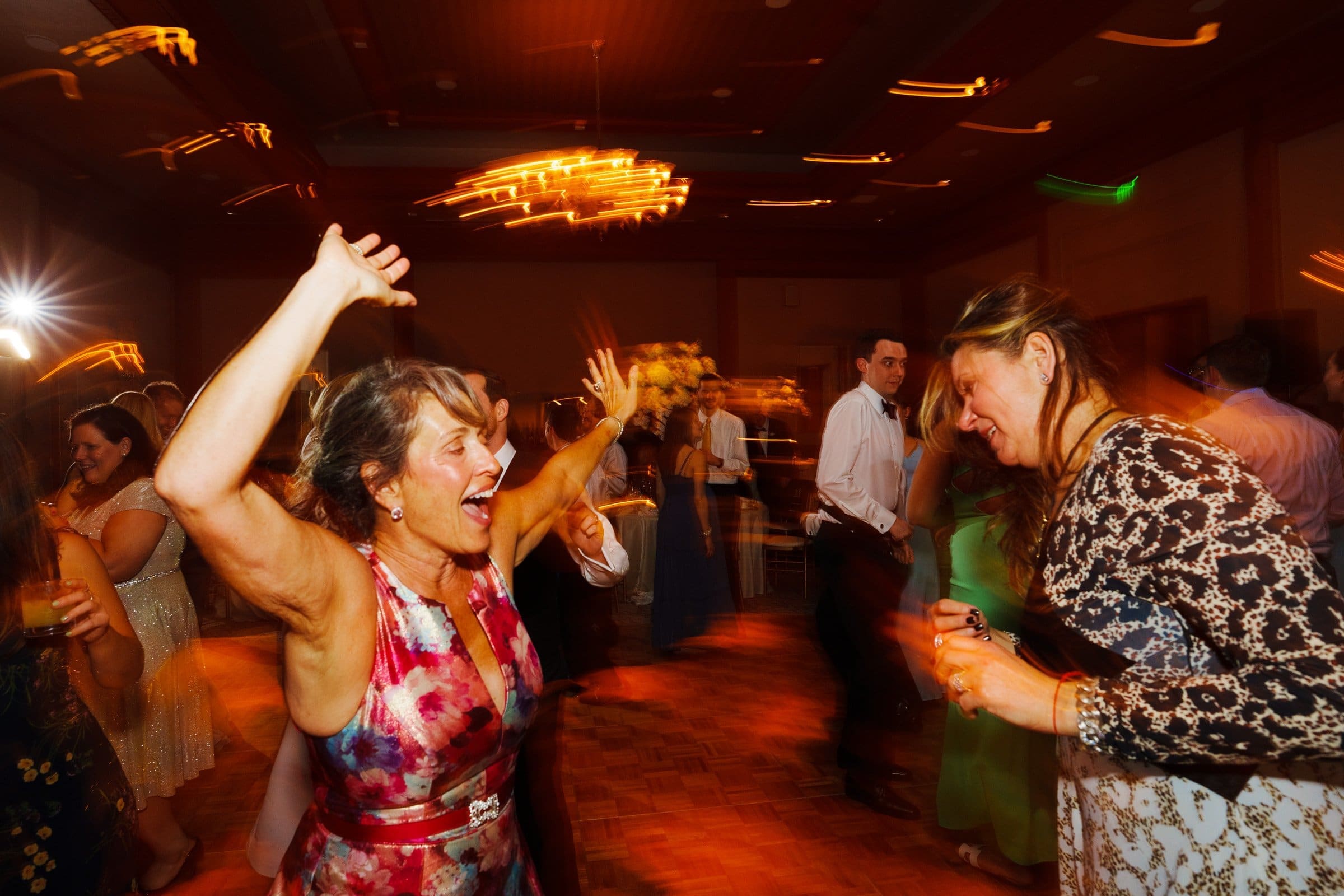 Two women joyfully dancing at a lively party. The woman on the left, reminiscent of a floral tapestry found in places like Duke Chapel, raises her arms, while the woman on the right in a patterned dress smiles warmly. Other guests sway in the background under warm, blurred lights akin to those at Umstead.