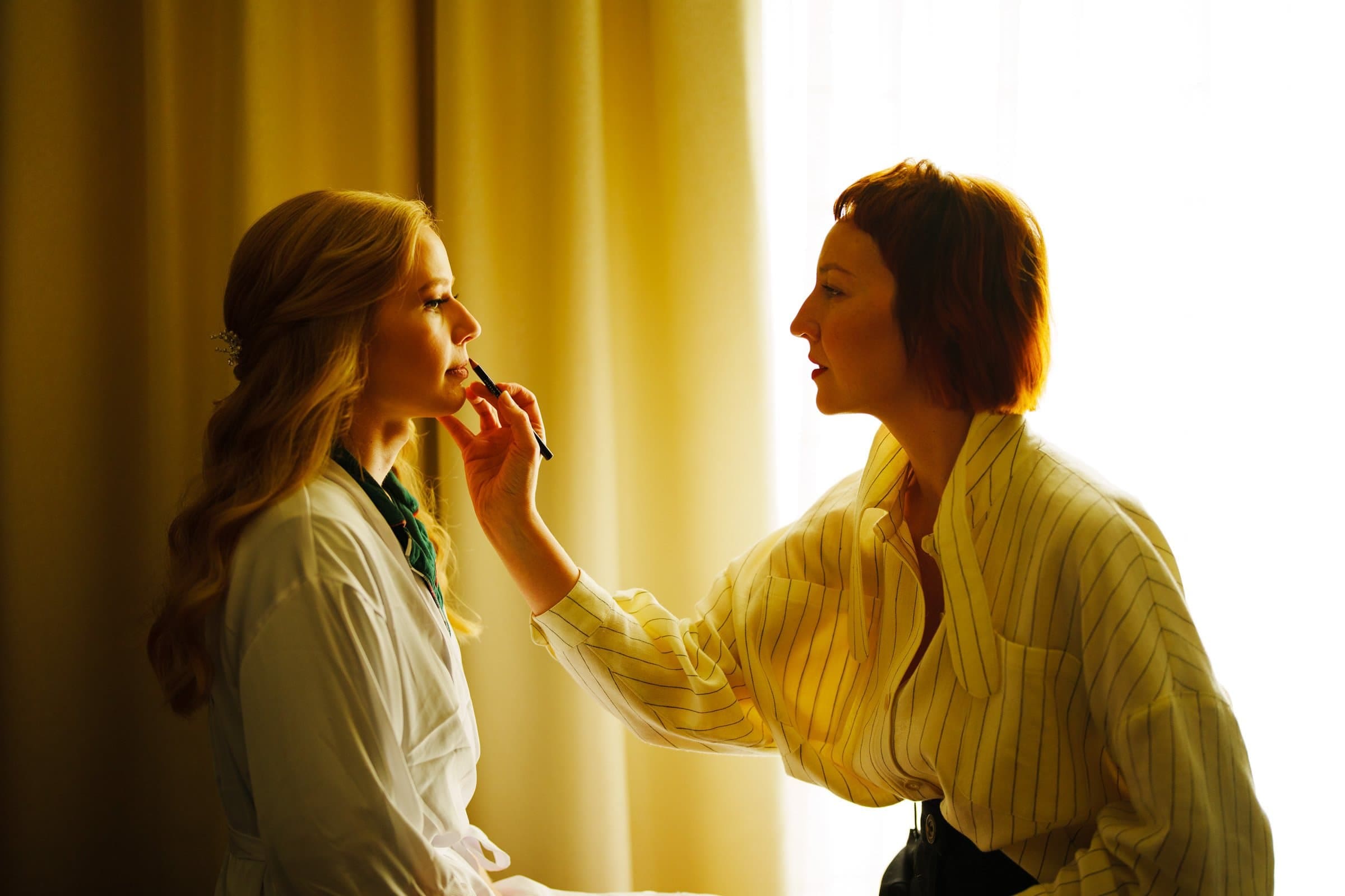 In a softly lit room reminiscent of a peaceful day at Umstead, a woman with long blonde hair sits in a robe. Another woman with short auburn hair delicately applies makeup to her face, while light filters gently through the drapes.
