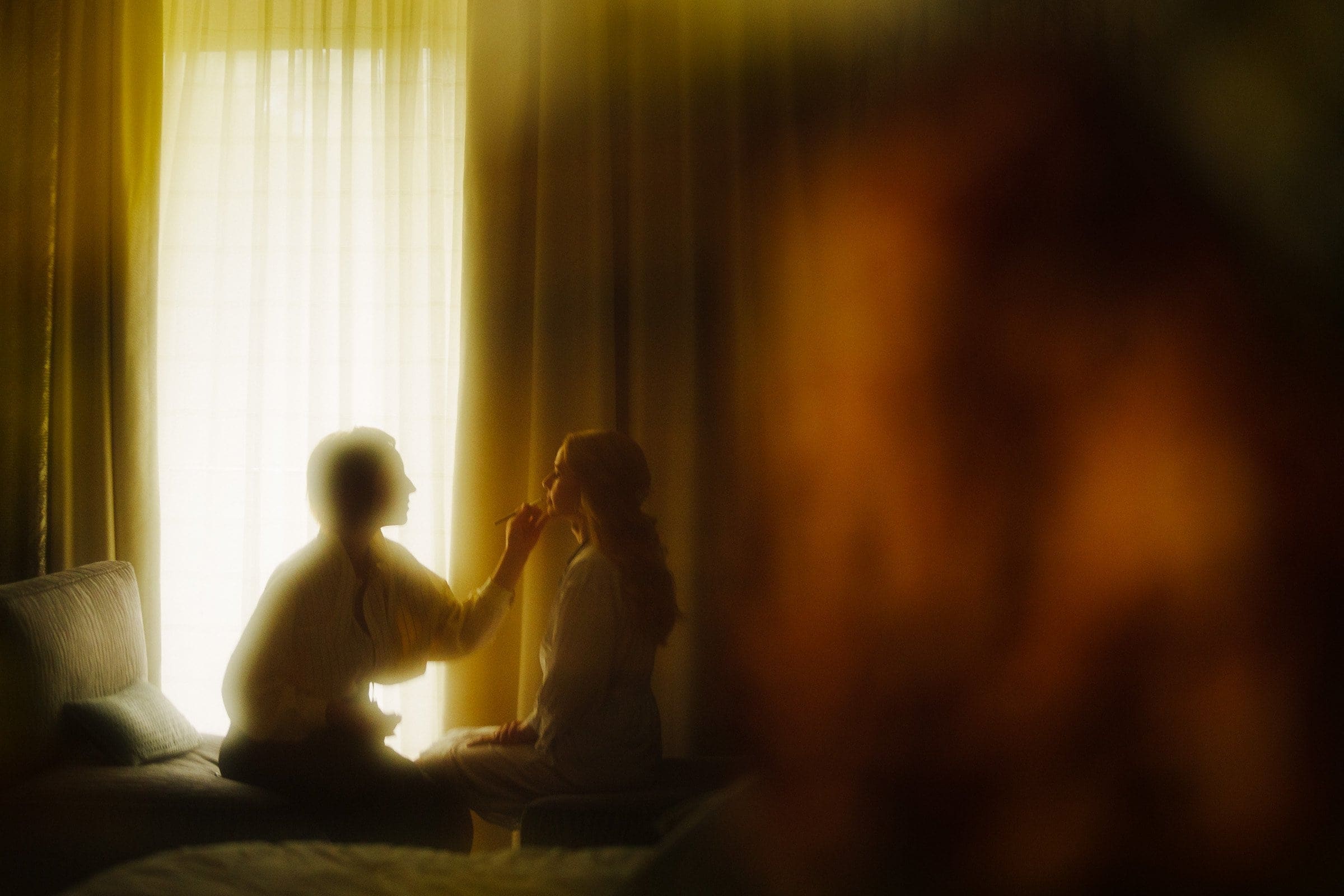 In the softly lit room, reminiscent of the tranquility found at Umstead, a makeup artist applies makeup to a seated woman in front of a warmly glowing window, creating an intimate and serene atmosphere.