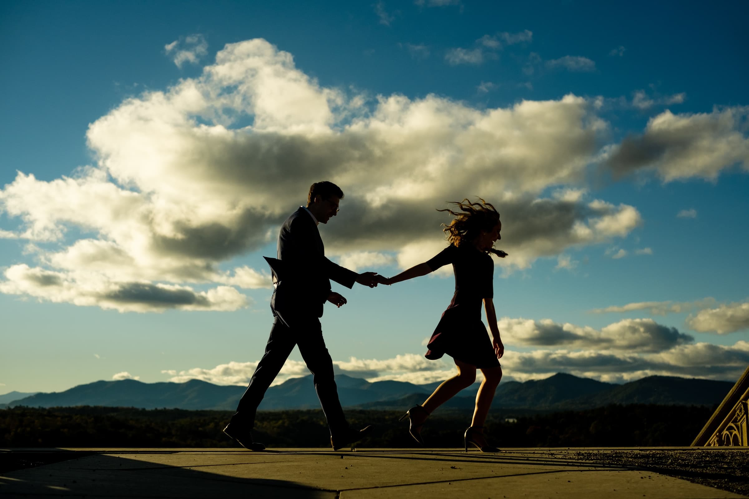 Silhouetted couple holding hands, running against a dramatic sky with fluffy clouds and mountains in the background—a perfect moment for Biltmore estate engagement photography.