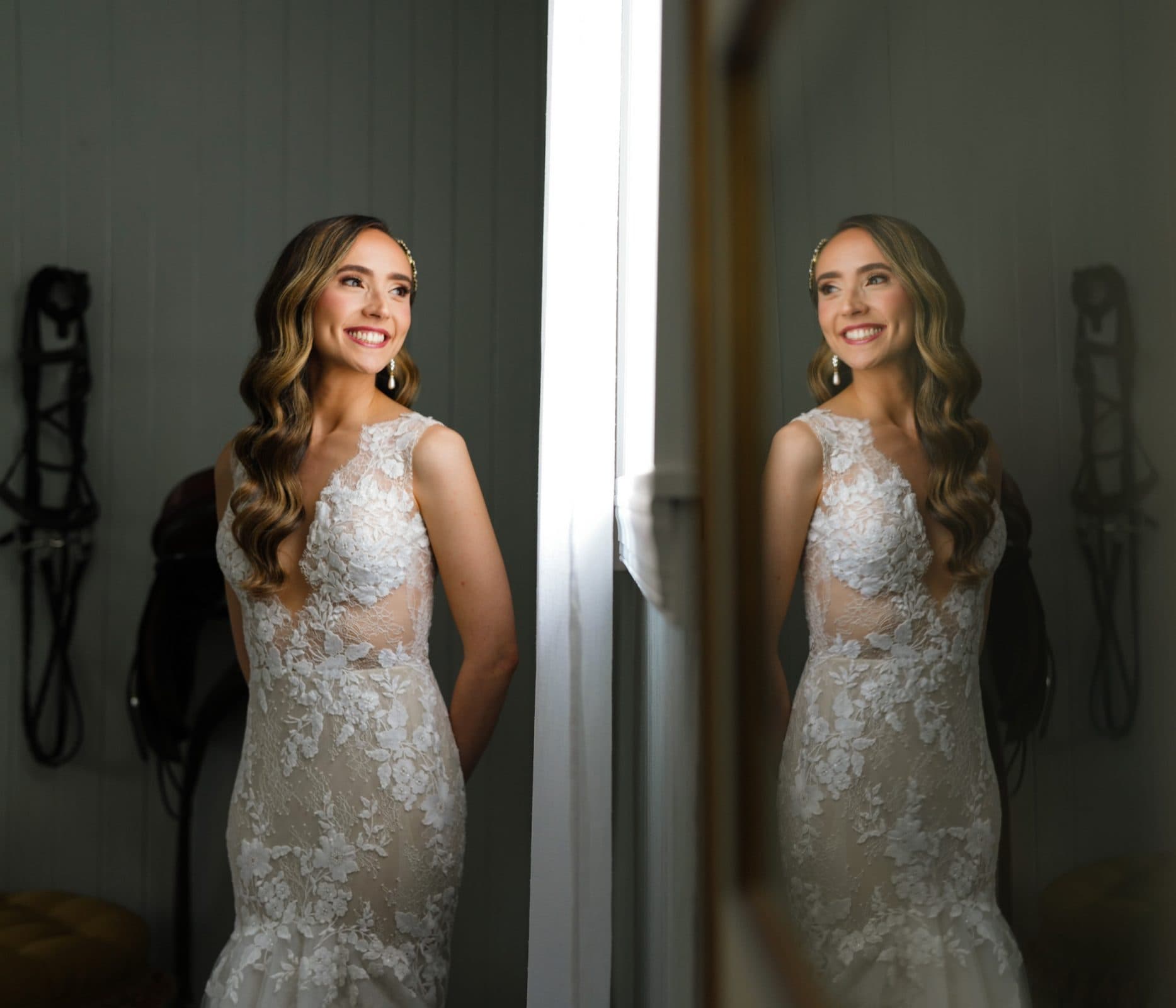 historic wakefield barn wedding photography - jennifer & stefan A woman in a lace wedding dress stands beside a mirror, smiling. Her reflection captures the intricate floral details of the gown. The background features equestrian gear hanging on the wall, adding a rustic touch to this Wakefield barn wedding scene.