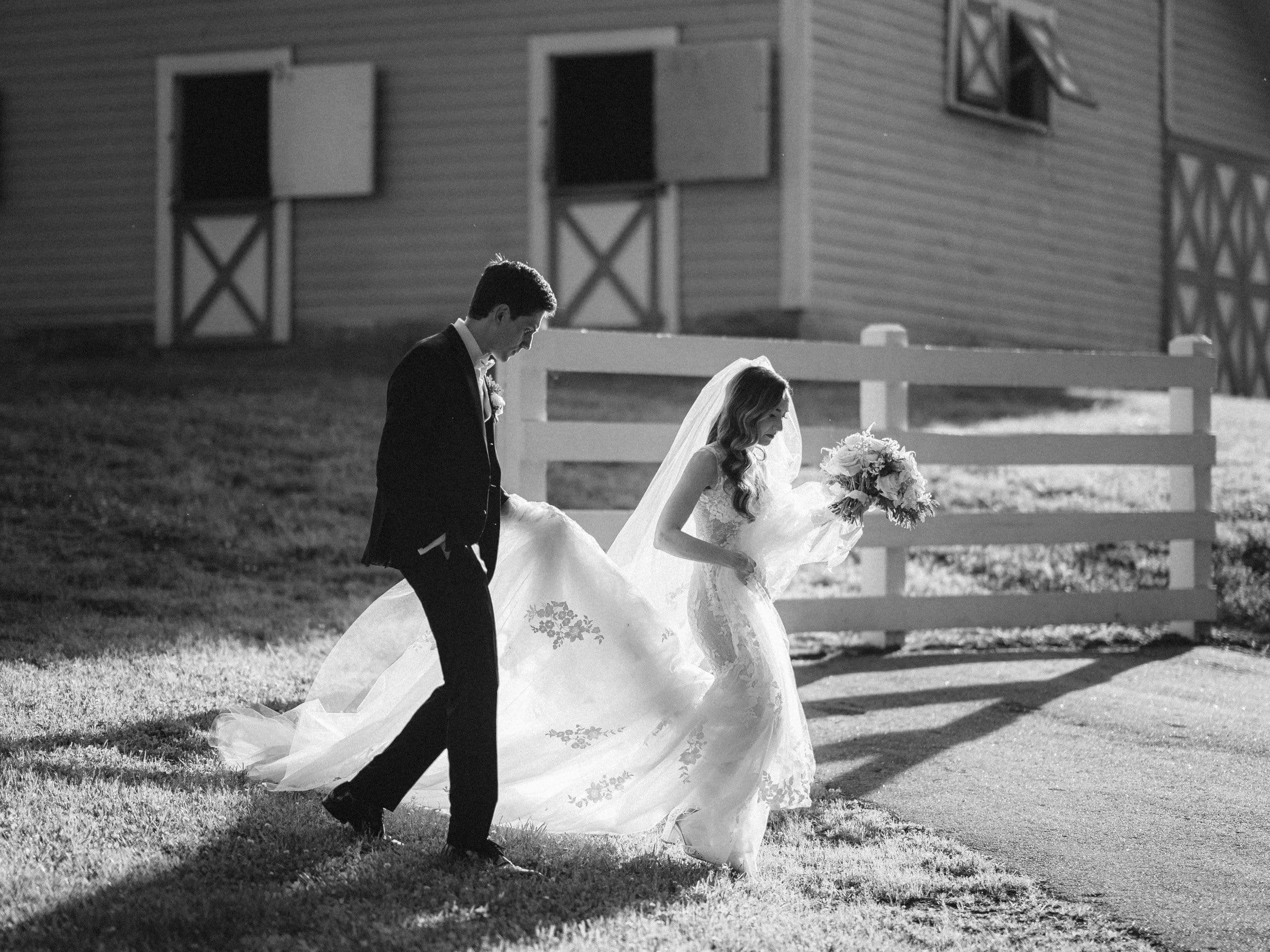 historic wakefield barn wedding photography - jennifer & stefan A bride and groom walk together in front of a barn. The bride holds a bouquet and wears a long, flowing veil. The groom wears a suit and pockets his hands. The scene is in black and white, with shadows cast on the ground.