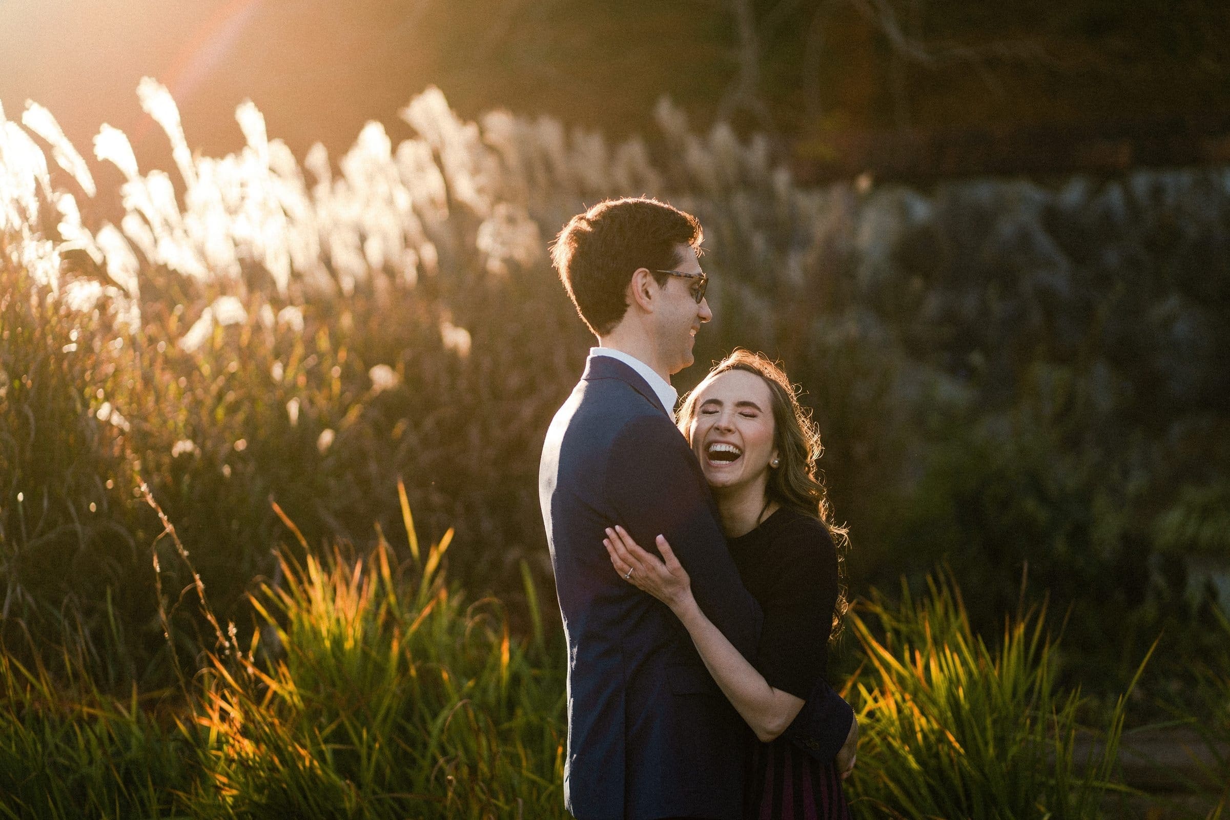 A couple embraces and laughs in a sunlit field at the Biltmore estate, surrounded by tall grass and brush. The sun filters through the scene, creating a warm, golden glow—perfect for engagement photography that captures their joy.