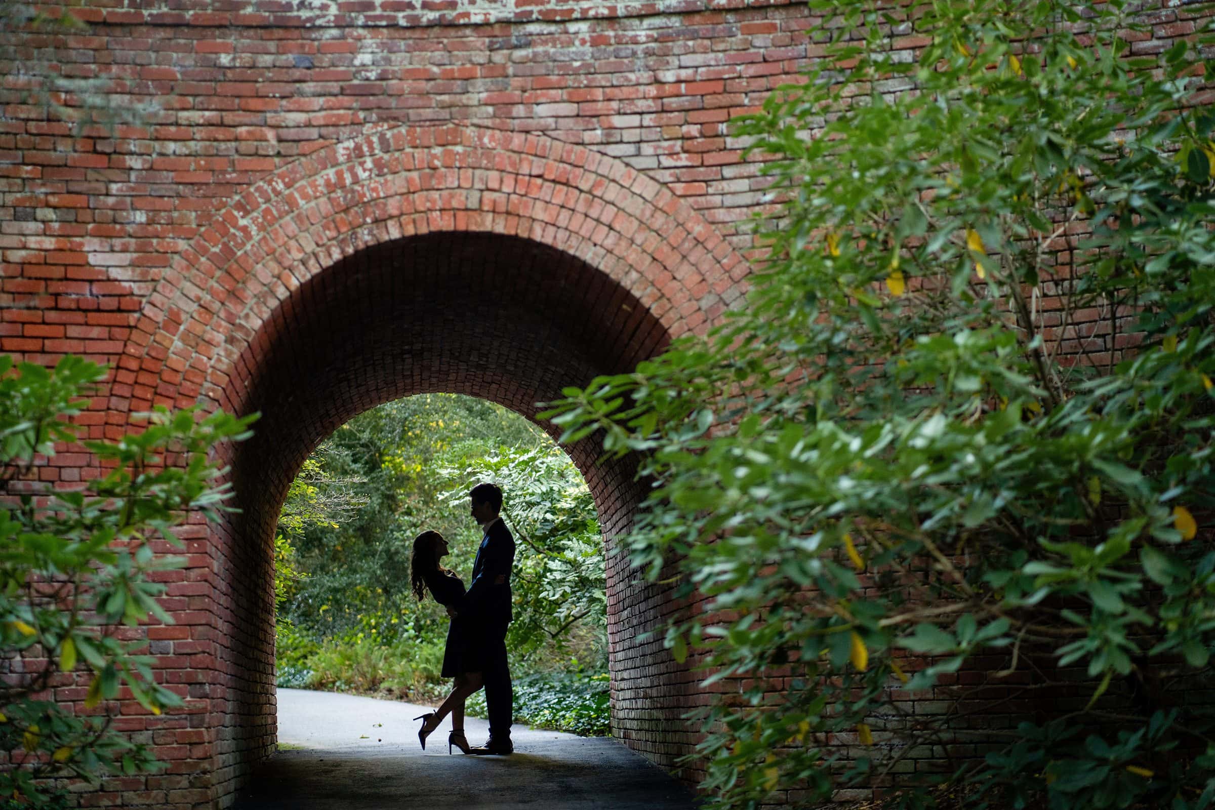 Silhouetted couple embracing under a brick archway, surrounded by lush greenery—a perfect moment of romance captured in Biltmore Estate engagement photography.