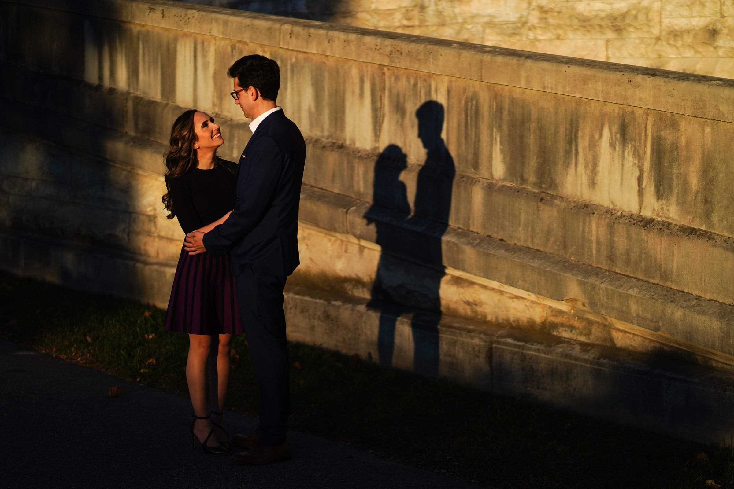 A couple stands closely together, gazing into each other's eyes at the Biltmore Estate, as their shadows are cast on a stone wall. The warm sunlight enhances the romantic atmosphere, capturing the essence of engagement photography.