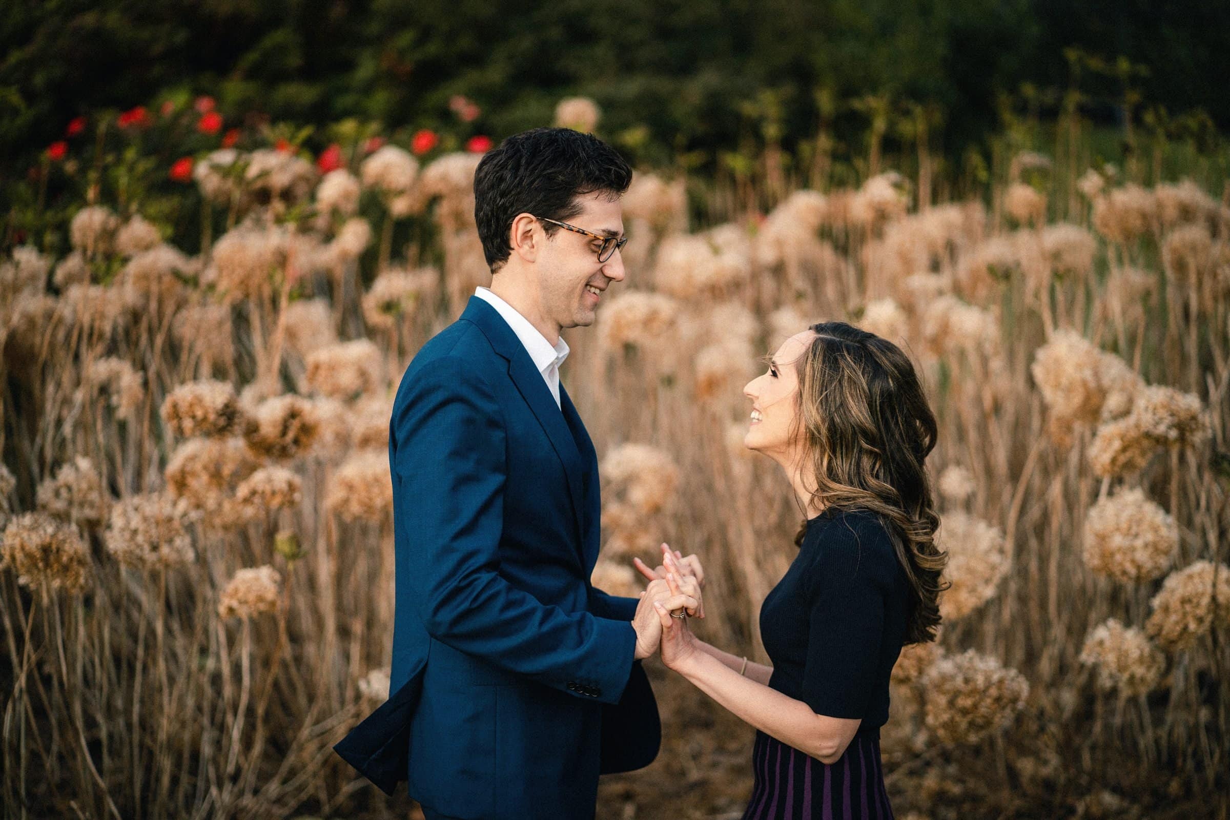 A couple stands facing each other, holding hands and smiling amid the enchanting scenery of the Biltmore Estate. The man, in a blue suit and glasses, complements the woman in her black top and dark skirt. Surrounded by tall dried flowers and lush green foliage, their engagement photography exudes charm.