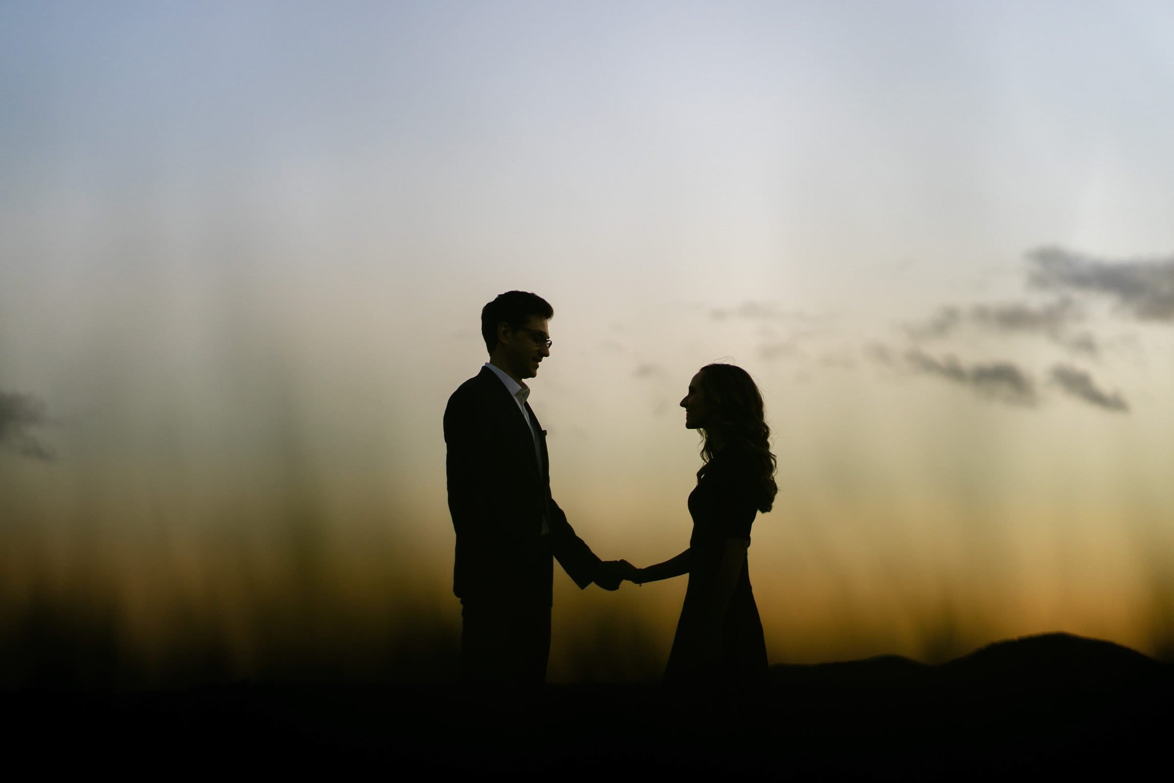 A silhouette of a couple holding hands against a sunset backdrop at the Biltmore Estate. The sky is a gradient of orange and blue with scattered clouds, creating a romantic and serene atmosphere perfect for engagement photography.