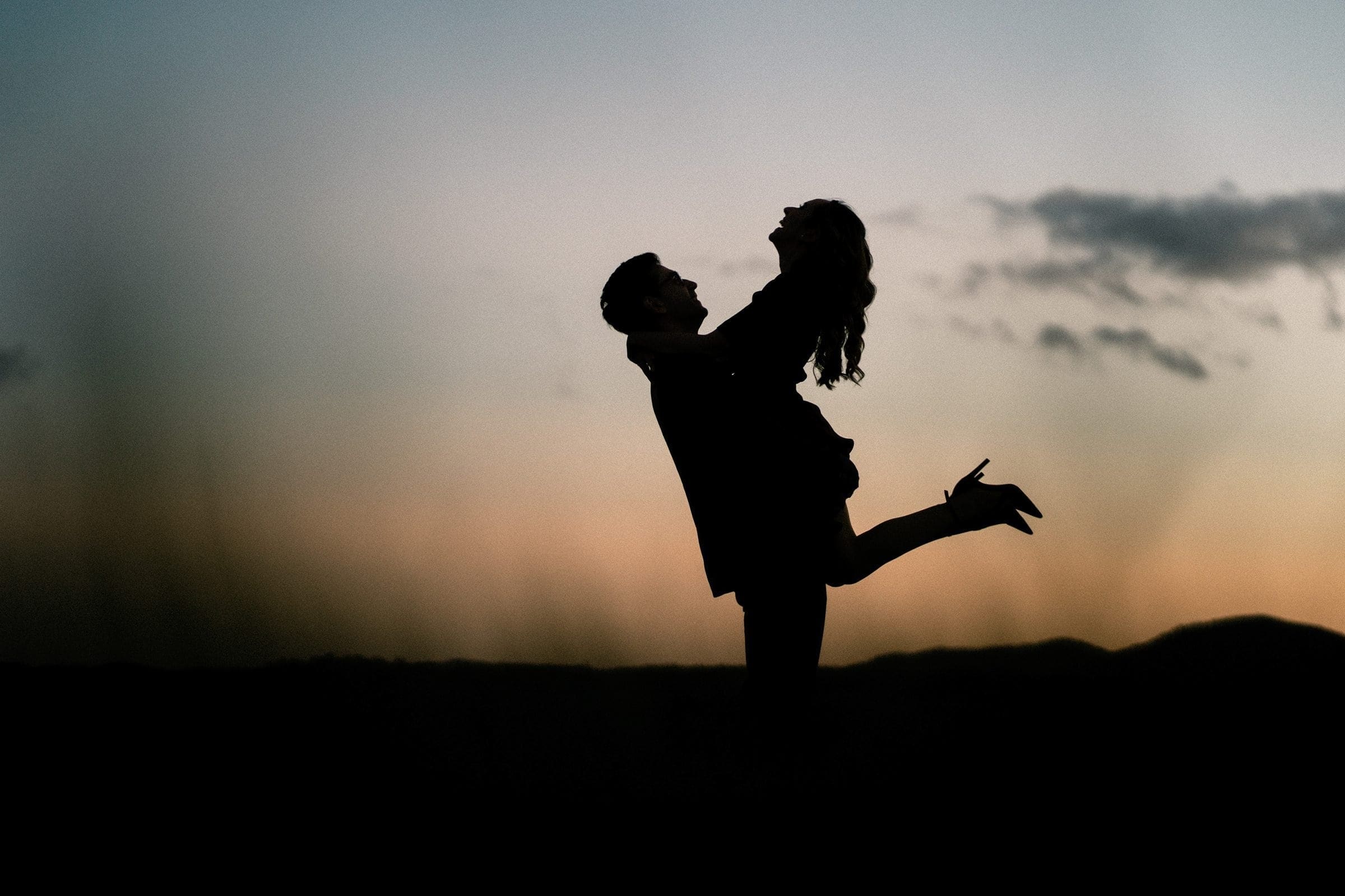 Silhouette of a couple against a sunset sky at the Biltmore Estate. The man lifts the joyful woman in an open landscape with soft hills in the background. The scene conveys a sense of romance and freedom, perfect for engagement photography.