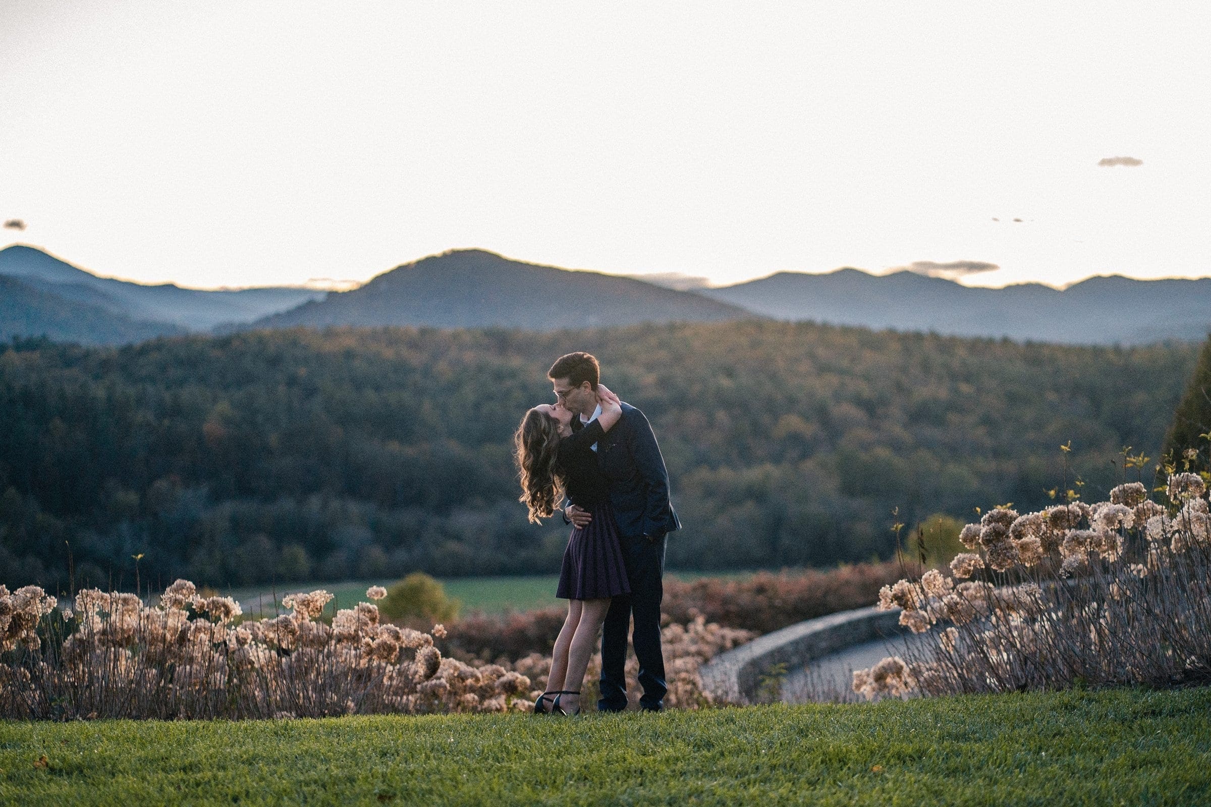 Amidst the serene, romantic atmosphere of an open field with mountains and a forest backdrop, a couple steals a kiss. The overcast sky adds to the charm as she graces a dark dress and he dons a suit, capturing their Biltmore estate engagement photography moment perfectly.