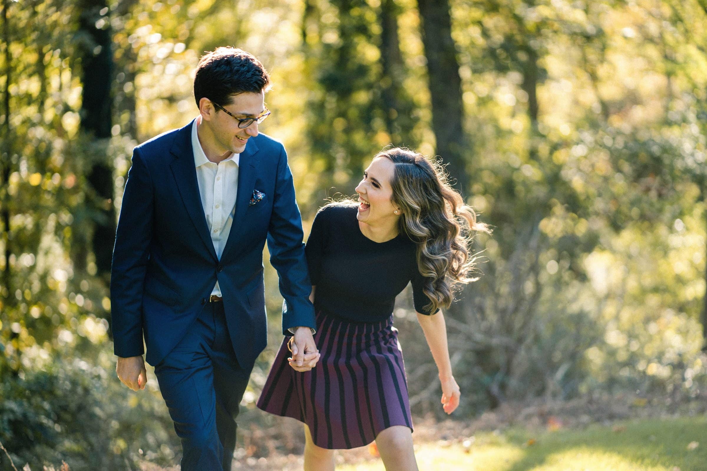 A couple joyfully walks hand in hand through the sunlit forest at the Biltmore estate. The man wears a dark suit, and the woman is in a black top and striped skirt. The trees' golden leaves create a warm, inviting atmosphere, perfect for engagement photography.