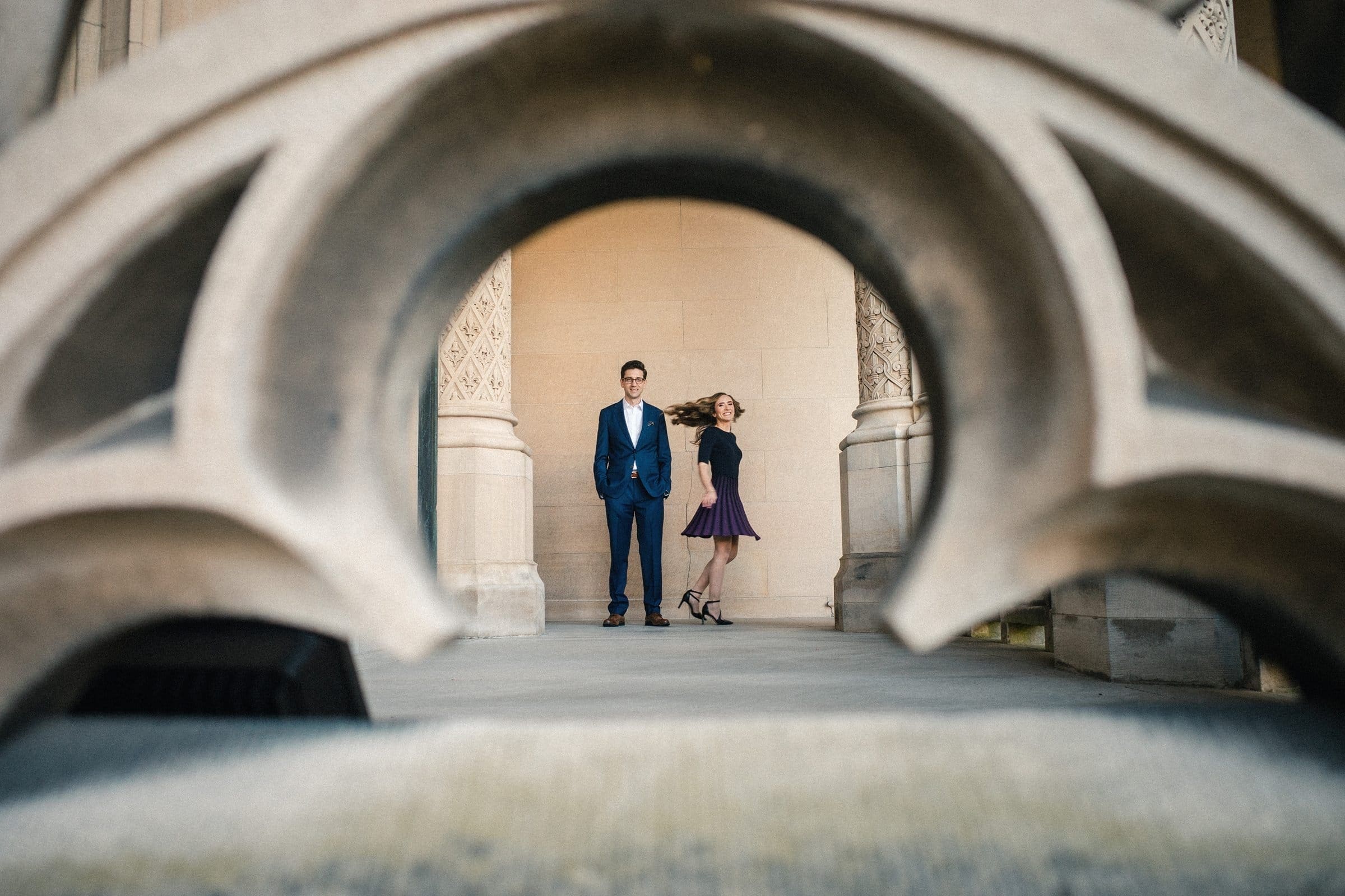 A man in a blue suit and a woman in a dark dress stand side by side in an architectural setting with ornate columns at the Biltmore Estate. The scene, perfect for engagement photography, is framed through an arched stone structure, adding a dramatic perspective.
