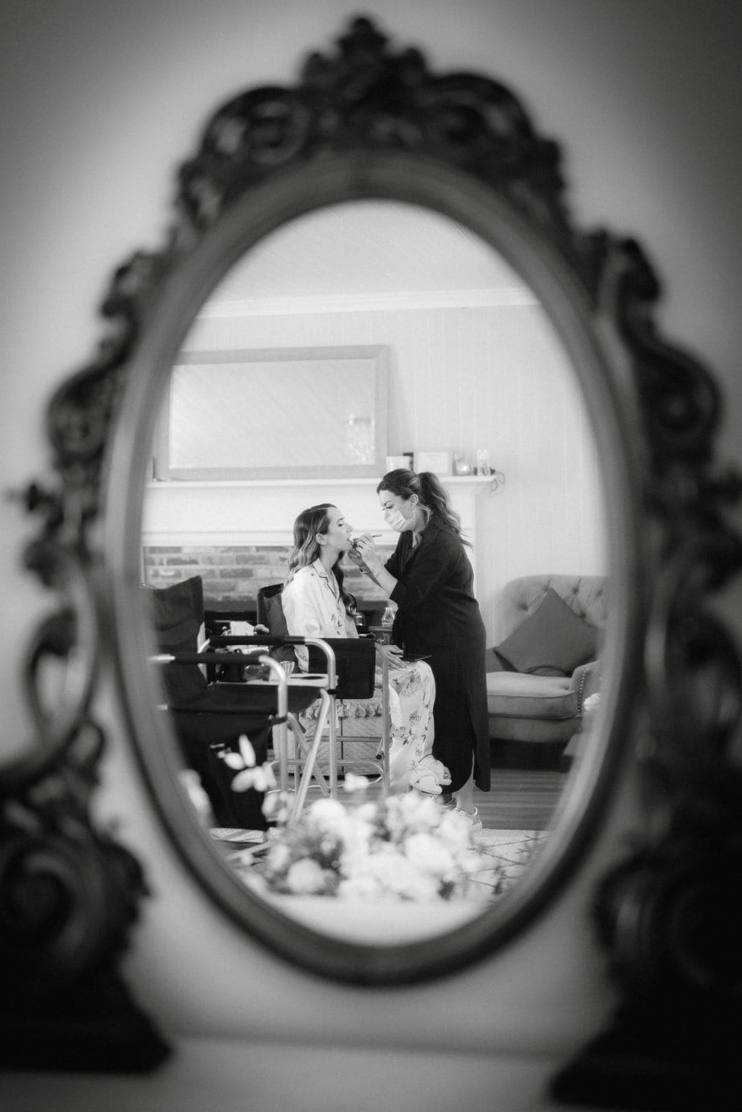 historic wakefield barn wedding photography - jennifer & stefan A black and white photo captures a woman at a Wakefield barn wedding, seated in front of a mirror as she gets her makeup done. The reflection reveals the elegant atmosphere with intricate mirror borders and the cozy setting of the barn in the background.