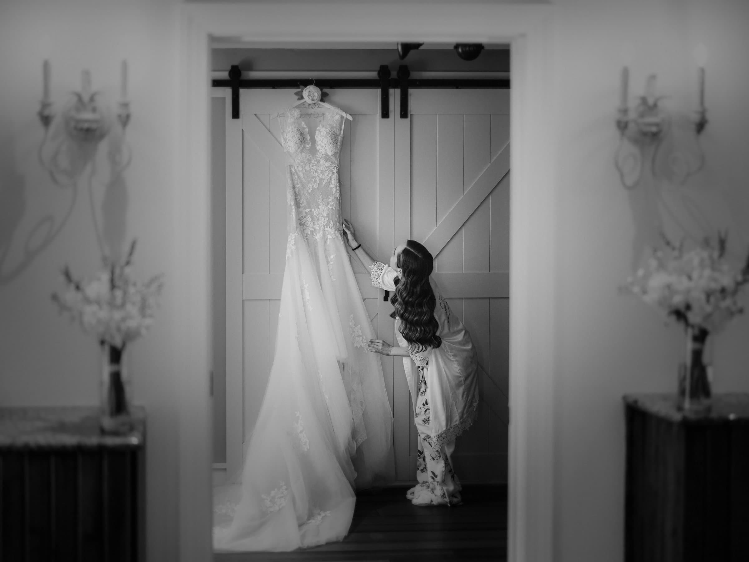 historic wakefield barn wedding photography - jennifer & stefan A bride admires her long, elegant wedding dress hanging on a sliding barn door at the Wakefield venue. The softly lit room, with decorative sconces and floral arrangements, captures the timeless essence of barn wedding photography in striking black and white.