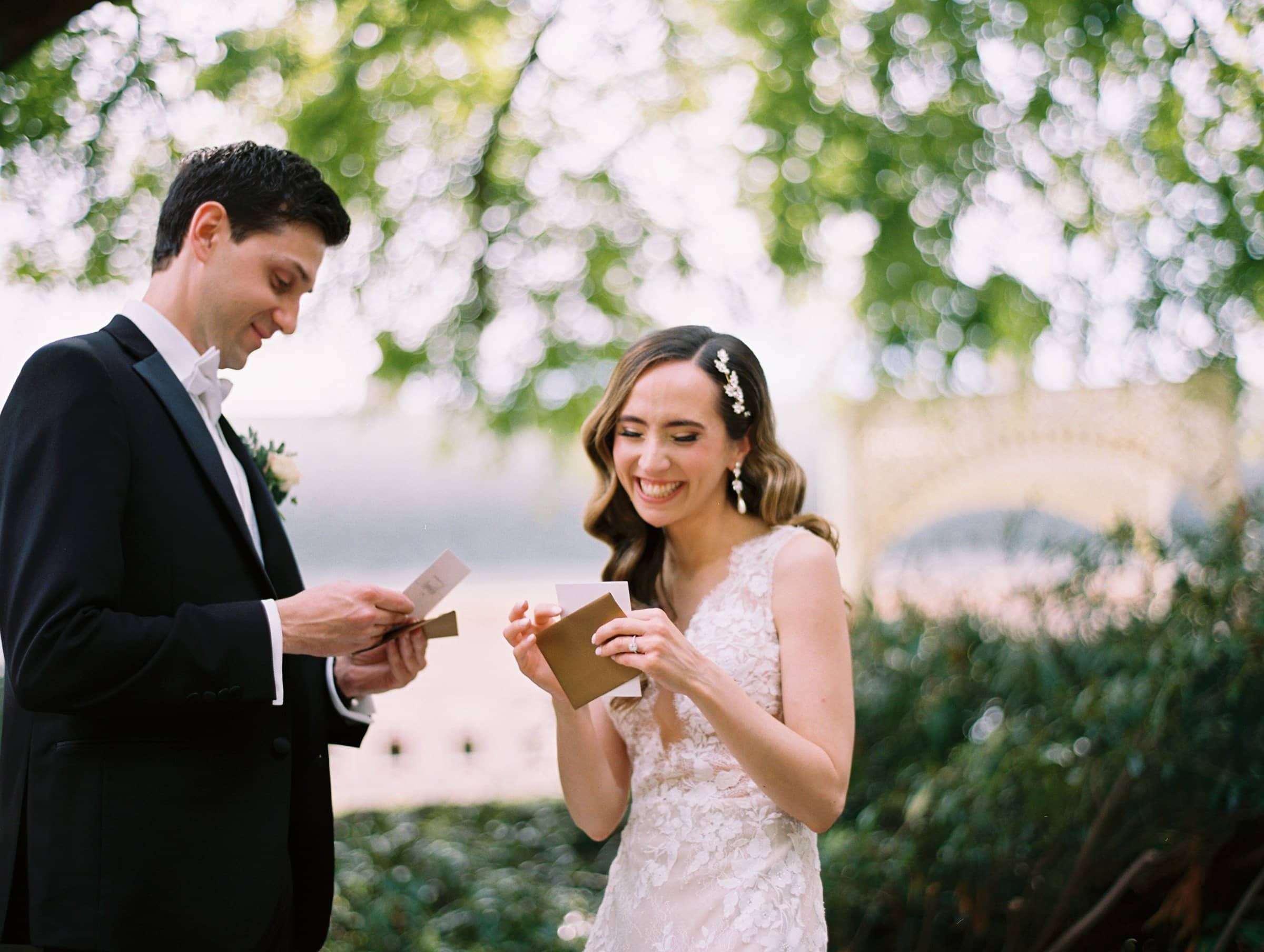 historic wakefield barn wedding photography - jennifer & stefan Amidst the lush greenery and soft, dappled sunlight of their Wakefield barn wedding, a bride in a lace dress laughs joyfully as she exchanges letters with her groom in a tuxedo, who grins broadly.