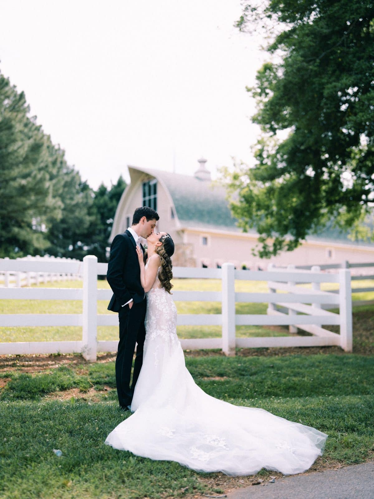 historic wakefield barn wedding photography - jennifer & stefan A bride and groom share a kiss in an outdoor setting captured by Wakefield barn wedding photography. The bride wears a white gown with a long train, and the groom is in a black tuxedo. They stand near a white fence with a barn and lush greenery in the background.
