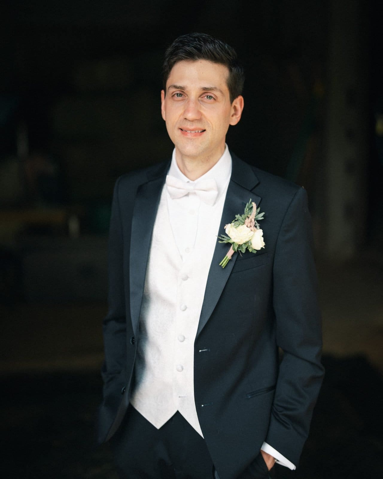 historic wakefield barn wedding photography - jennifer & stefan A man in a black tuxedo with a white bow tie and vest stands confidently, embodying elegance at a Wakefield barn wedding. A small boutonniere graces his jacket as he places his hand in his pocket, while the softly blurred background highlights his serene expression.