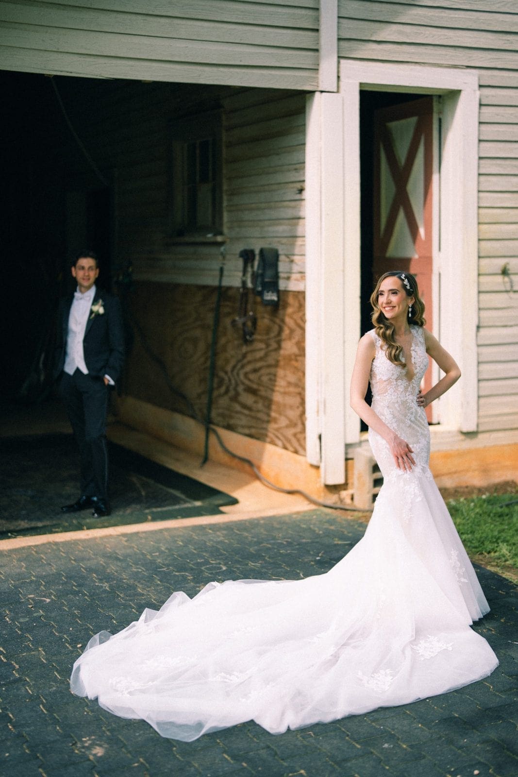 historic wakefield barn wedding photography - jennifer & stefan At Wakefield, a bride in a white gown poses gracefully before a wooden barn, her long train elegantly spread across the ground. The groom stands nearby, captured in perfect harmony by wedding photography as he gazes lovingly from the barn entrance.