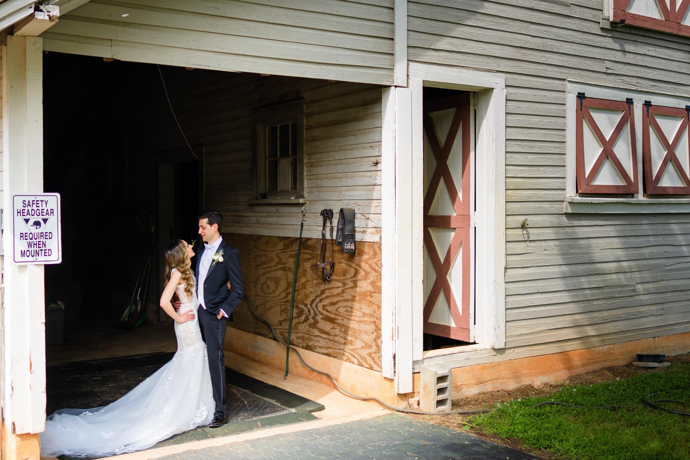 historic wakefield barn wedding photography - jennifer & stefan A bride and groom stand smiling, facing each other at a Wakefield barn wedding. The bride wears a long white gown, and the groom is in a dark suit. Sunlight bathes the rustic wooden barn's textured walls and open doors, capturing the perfect moment for wedding photography.