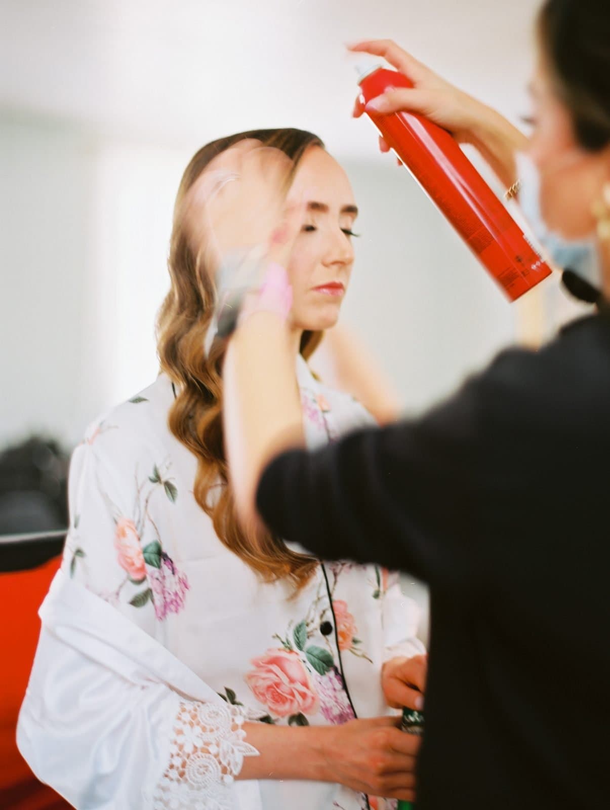 historic wakefield barn wedding photography - jennifer & stefan A woman with her eyes closed is having her hair styled with hairspray by another person, perfectly capturing a tranquil moment of preparation. She wears a white floral robe, evoking elegance. The softly blurred background hints at an indoor setting, perhaps during a Wakefield barn wedding photography session.