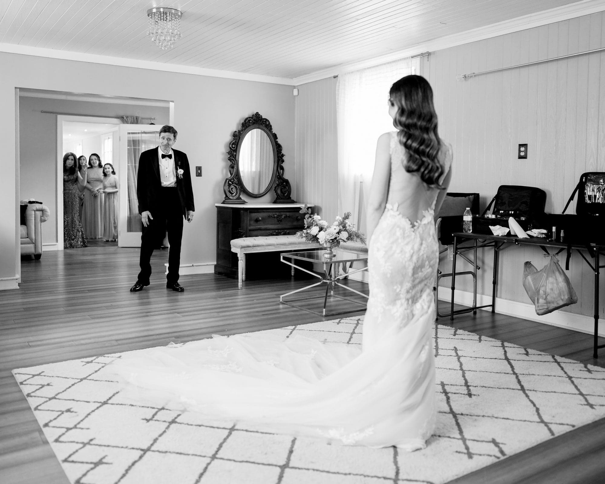 historic wakefield barn wedding photography - jennifer & stefan A bride in a flowing gown stands on the carpeted floor of Wakefield Barn, facing an older man in a suit, who looks surprised and delighted. In the background, several women in dresses watch from a doorway. The setting is elegantly lit, capturing the perfect wedding photography moment.