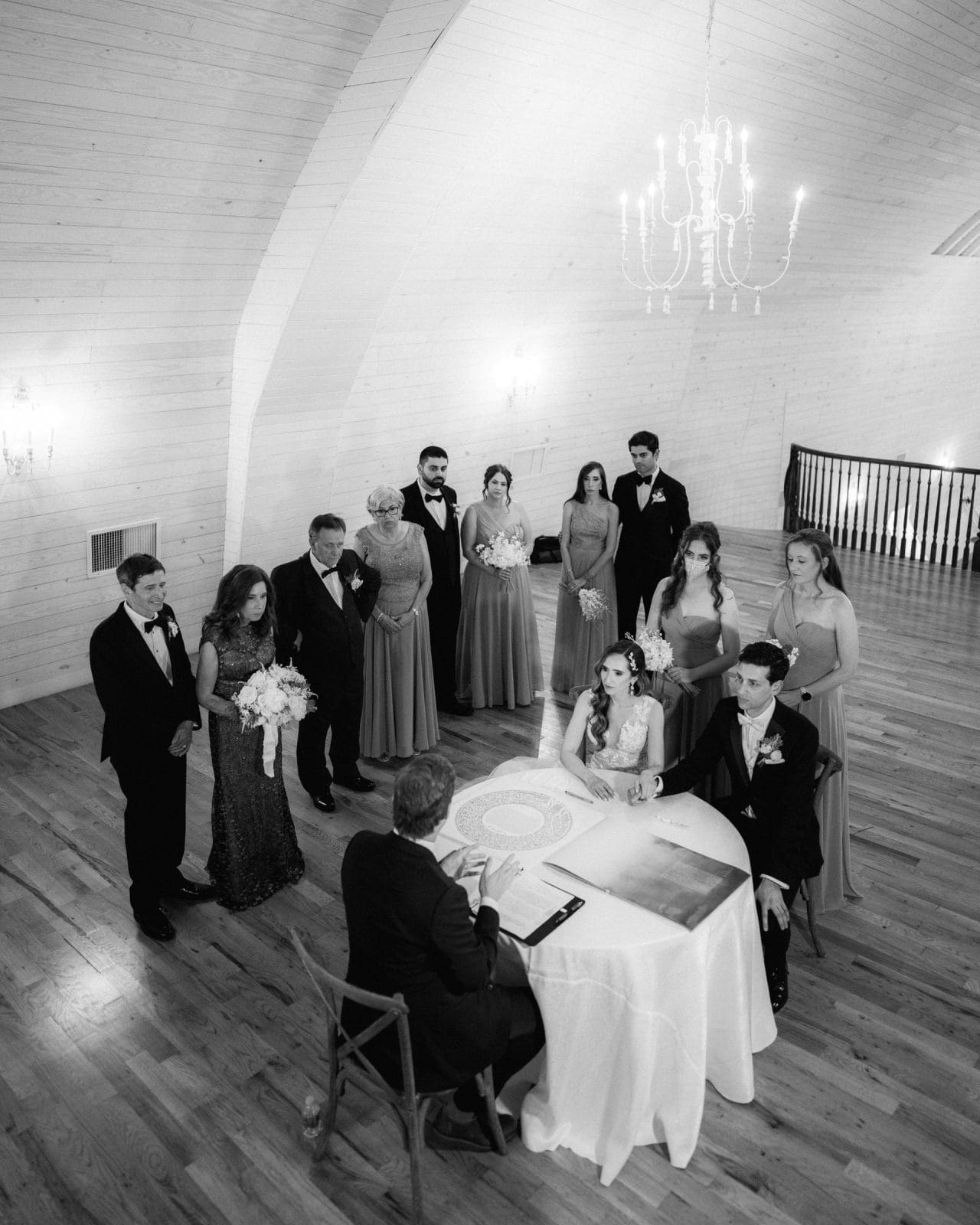 historic wakefield barn wedding photography - jennifer & stefan A black and white photo captures a Wakefield barn wedding ceremony in a large room with chandeliers. A couple sits at a table with a person officiating, while their wedding party stands around them. The bridesmaids wear long dresses, and the groomsmen are in suits.
