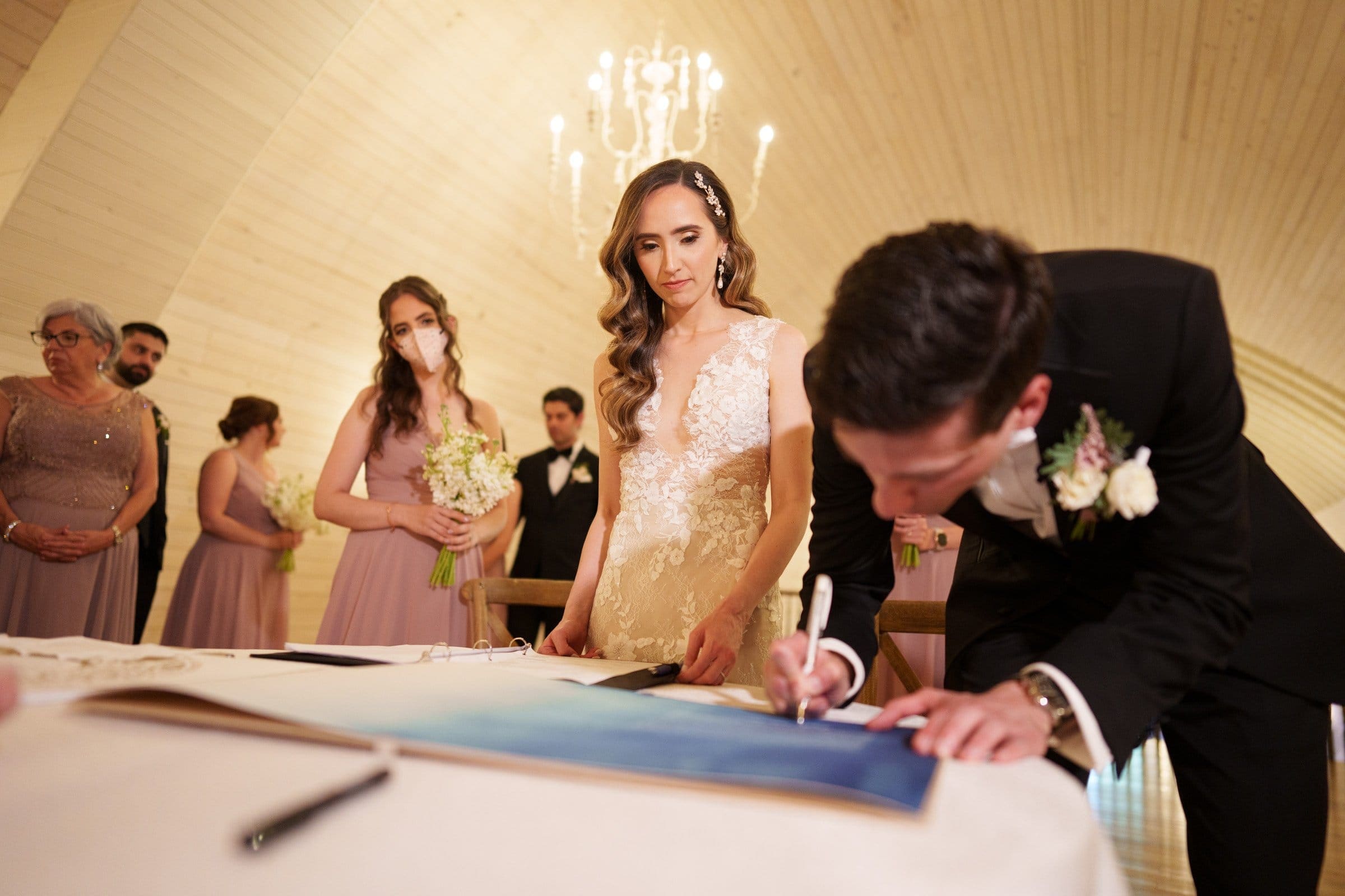 historic wakefield barn wedding photography - jennifer & stefan A bride in a lace gown observes as a groom signs a document on the table at their Wakefield Barn wedding. Bridesmaids in matching dresses and guests in formal attire mingle in the elegantly lit room, with chandeliers casting a warm glow over the scene.