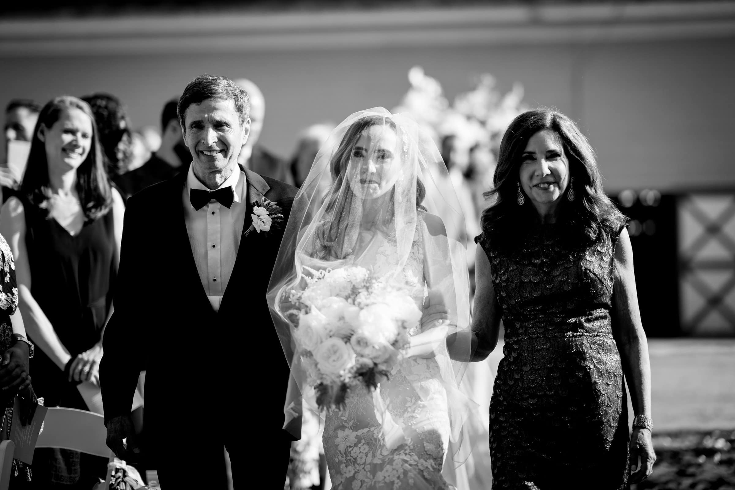 historic wakefield barn wedding photography - jennifer & stefan A black and white photo captures a timeless moment at a Wakefield barn wedding. The bride gracefully walks down the aisle, holding a bouquet and wearing a veil. With her are two escorts: a man in a tuxedo on her left and a woman in lace on her right, while guests seated on either side smile warmly.