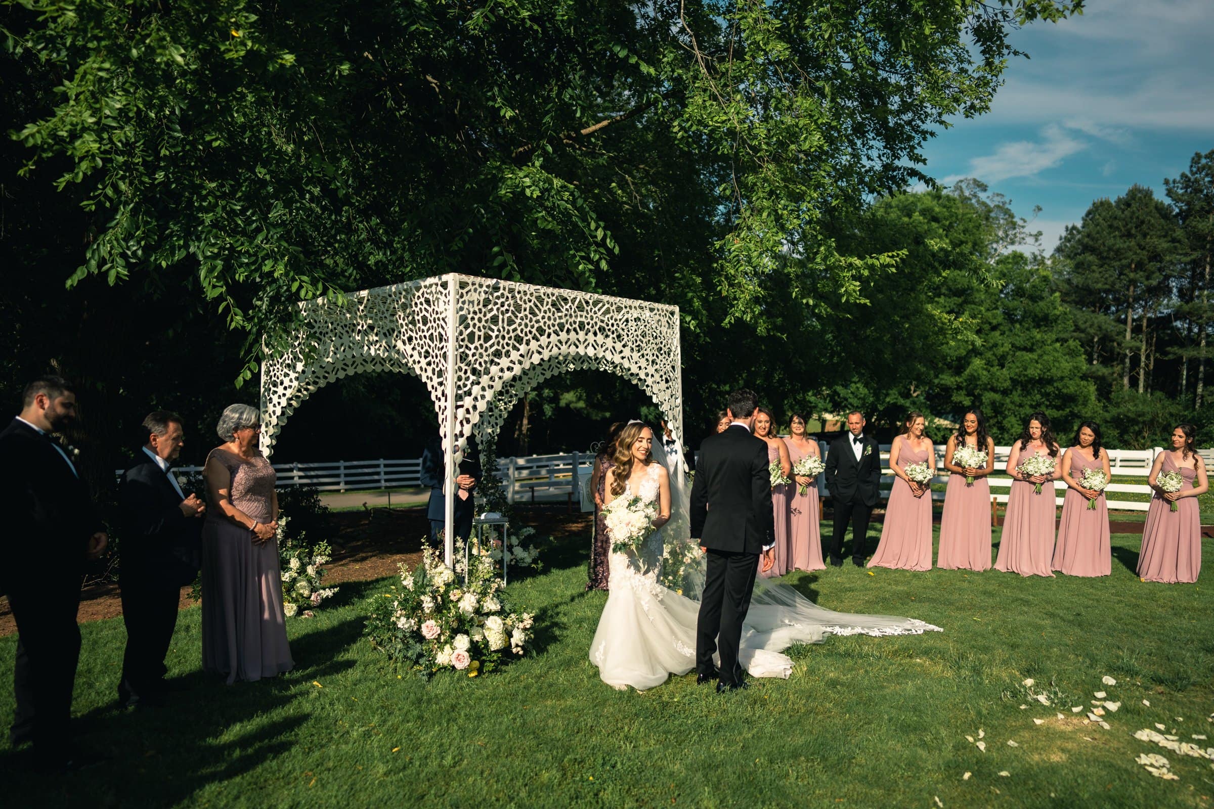 historic wakefield barn wedding photography - jennifer & stefan An outdoor wedding ceremony with a bride and groom under a floral arch is beautifully captured by Wakefield Barn Wedding Photography. Bridesmaids in pink dresses and groomsmen in dark suits stand nearby, set against a grassy lawn with lush greenery under a sunny blue sky.