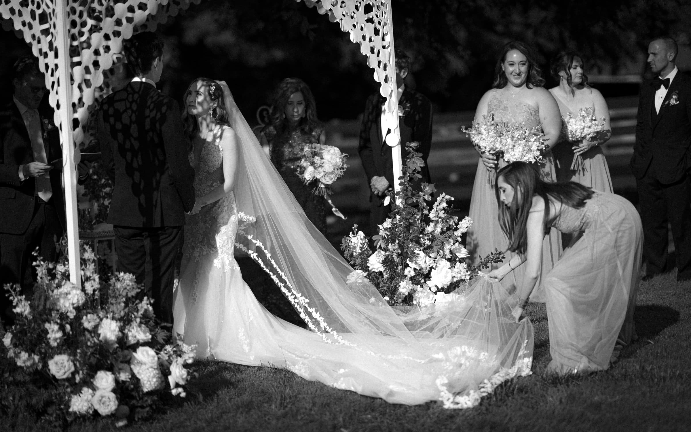 historic wakefield barn wedding photography - jennifer & stefan A black and white photo of a Wakefield barn wedding ceremony captures the bride, under an ornate archway, smiling at the groom. A bridesmaid adjusts her long veil as others stand nearby with bouquets, watching the couple.